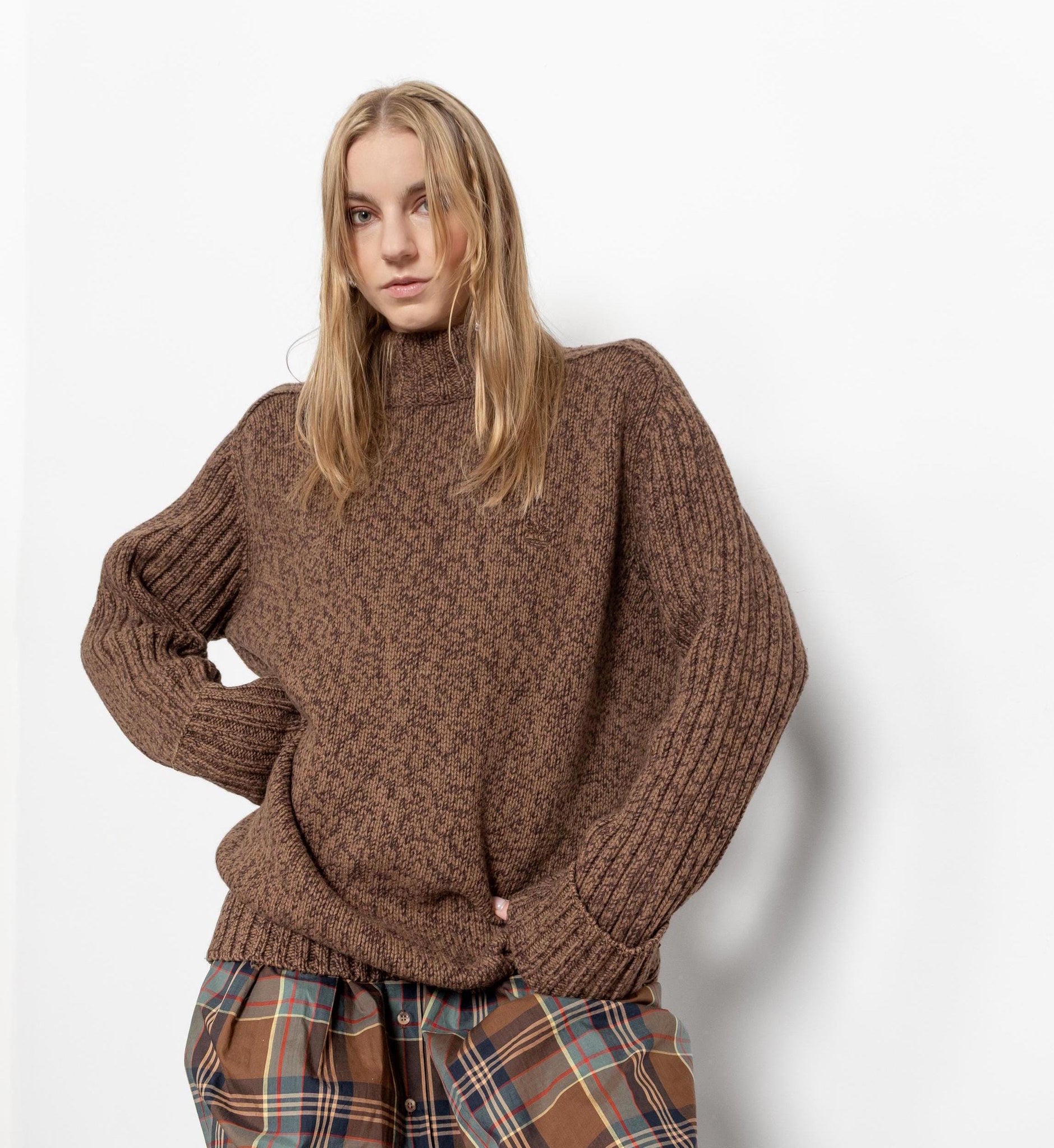 A female model wearing a heather brown wool turtleneck sweater with oversized fit and ribbed sleeves. She is also wearing patterned pants.