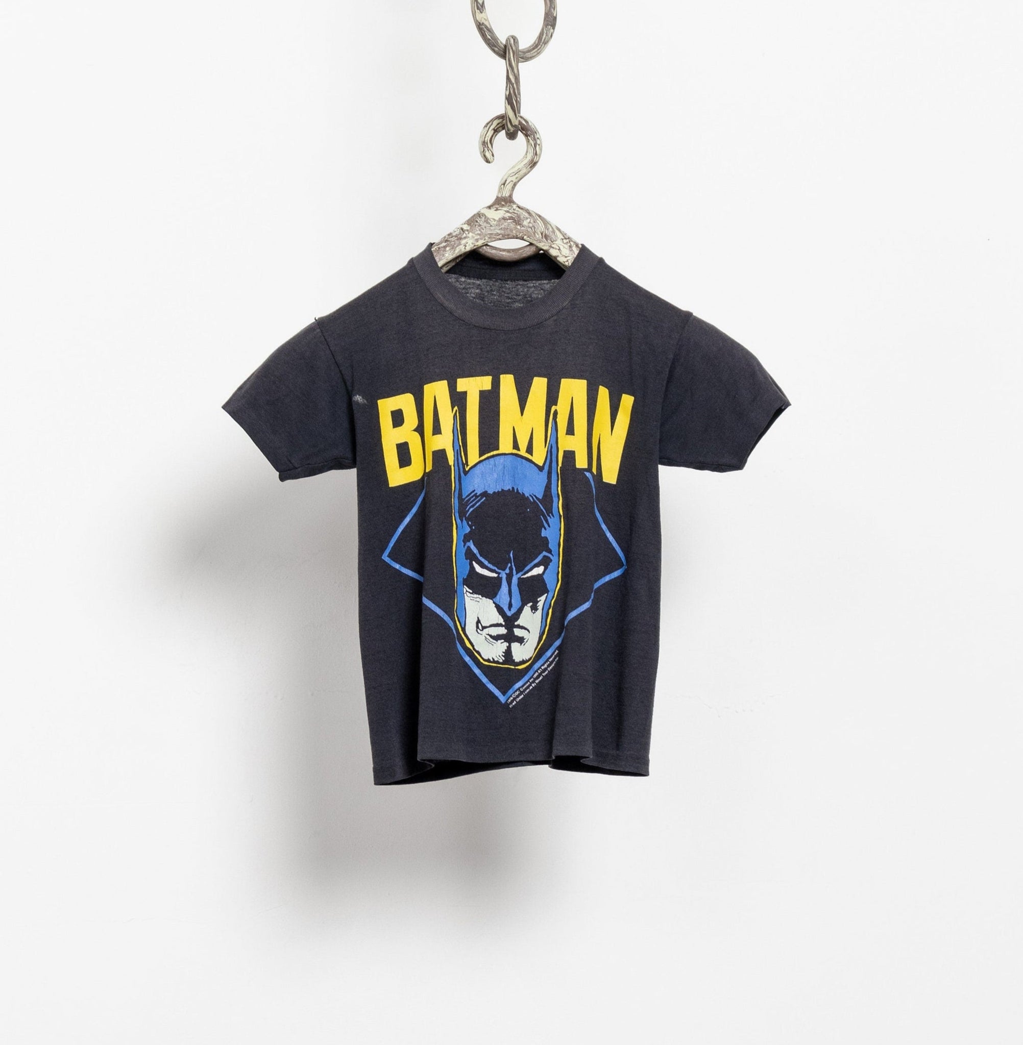 A vintage official Batman small fit t-shirt with a graphic print of the Batman character in blue and yellow on a dark background, displayed on a hanger.