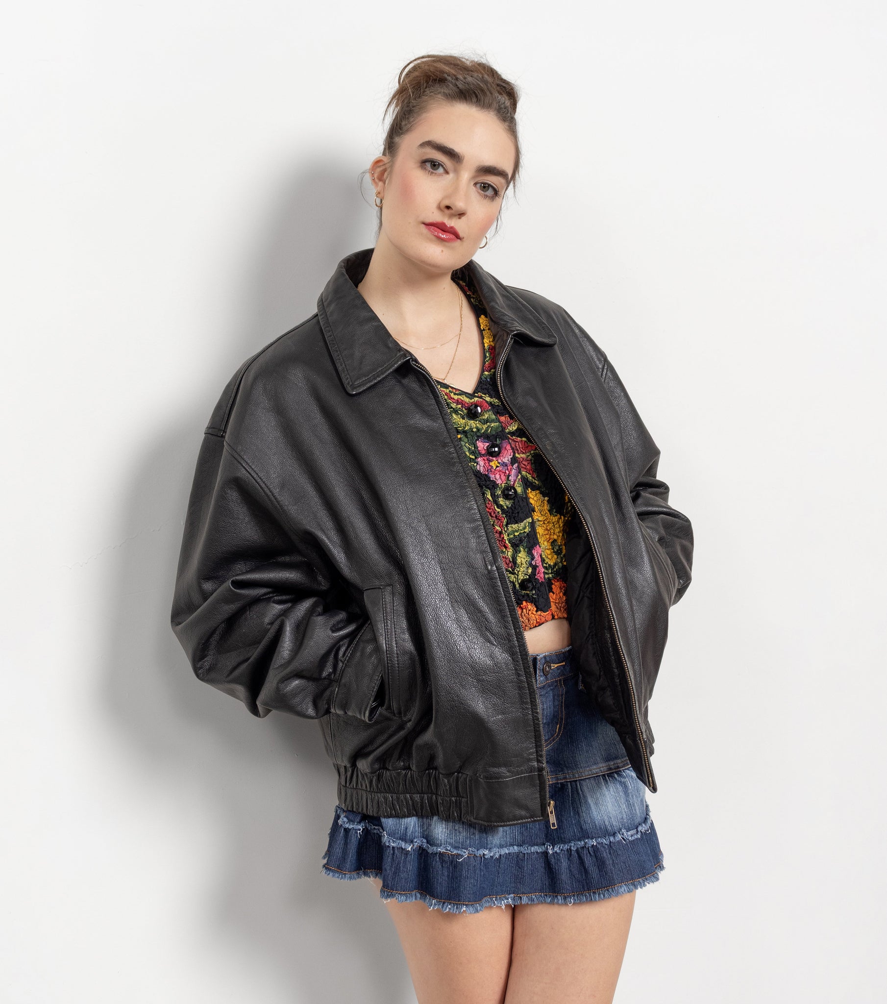 Woman wearing a black leather jacket over a colorful top and denim skirt on a plain background
