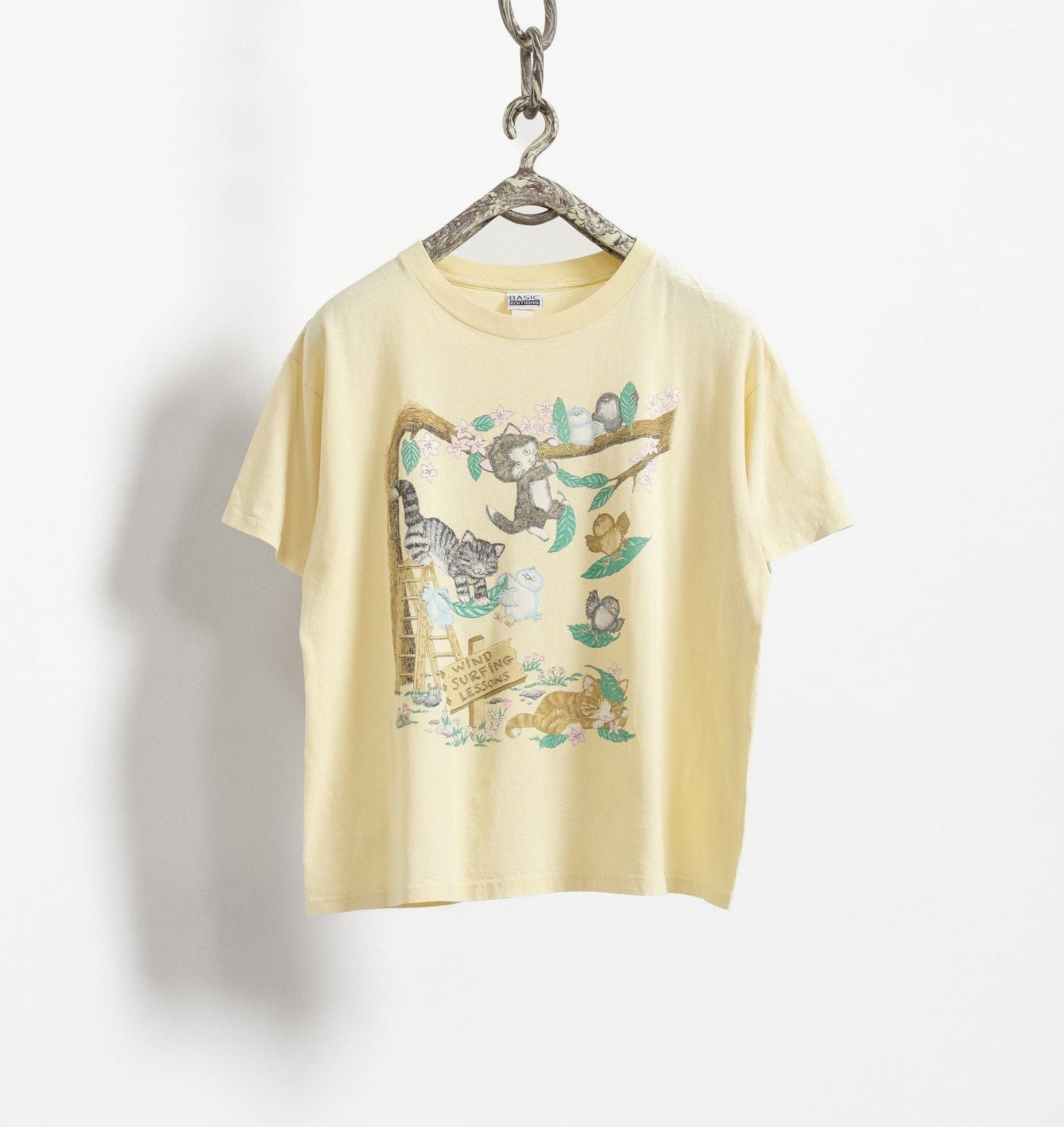 A vintage yellow t-shirt with a graphic of cats and birds on it, hanging on a display hanger against a white background.