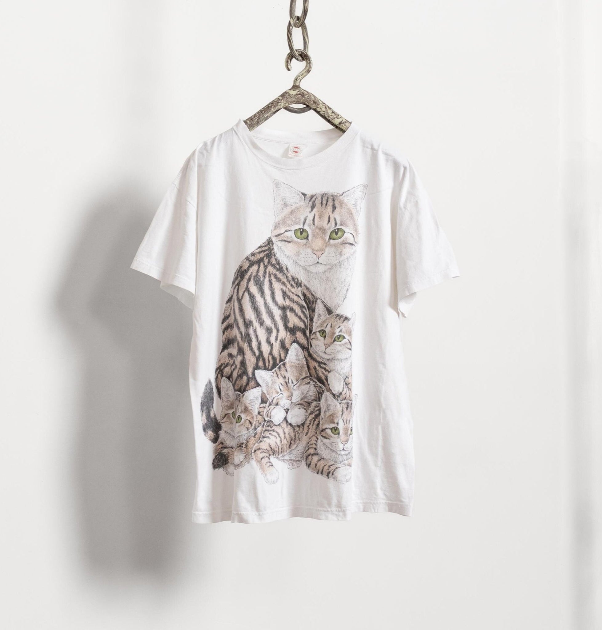 A white vintage t-shirt with a graphic of a mother cat and her kittens, hanging on a hanger against a white background.