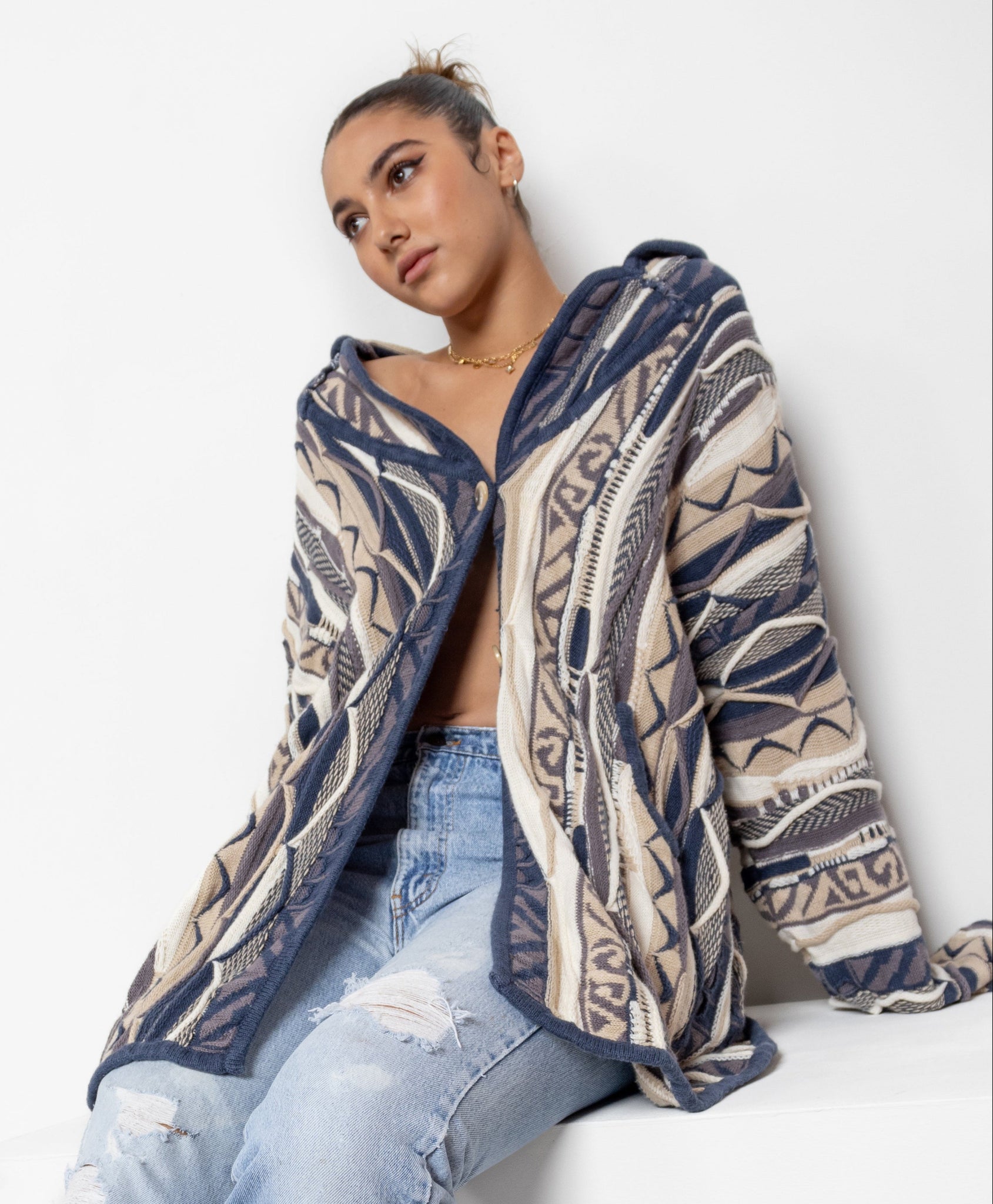 A woman sitting against a white background wearing a vintage 90's patterned 100% cotton knit dad cardigan in hues of blue, brown, beige, cream, and grey, with big tortishell buttons and an oversized fit.