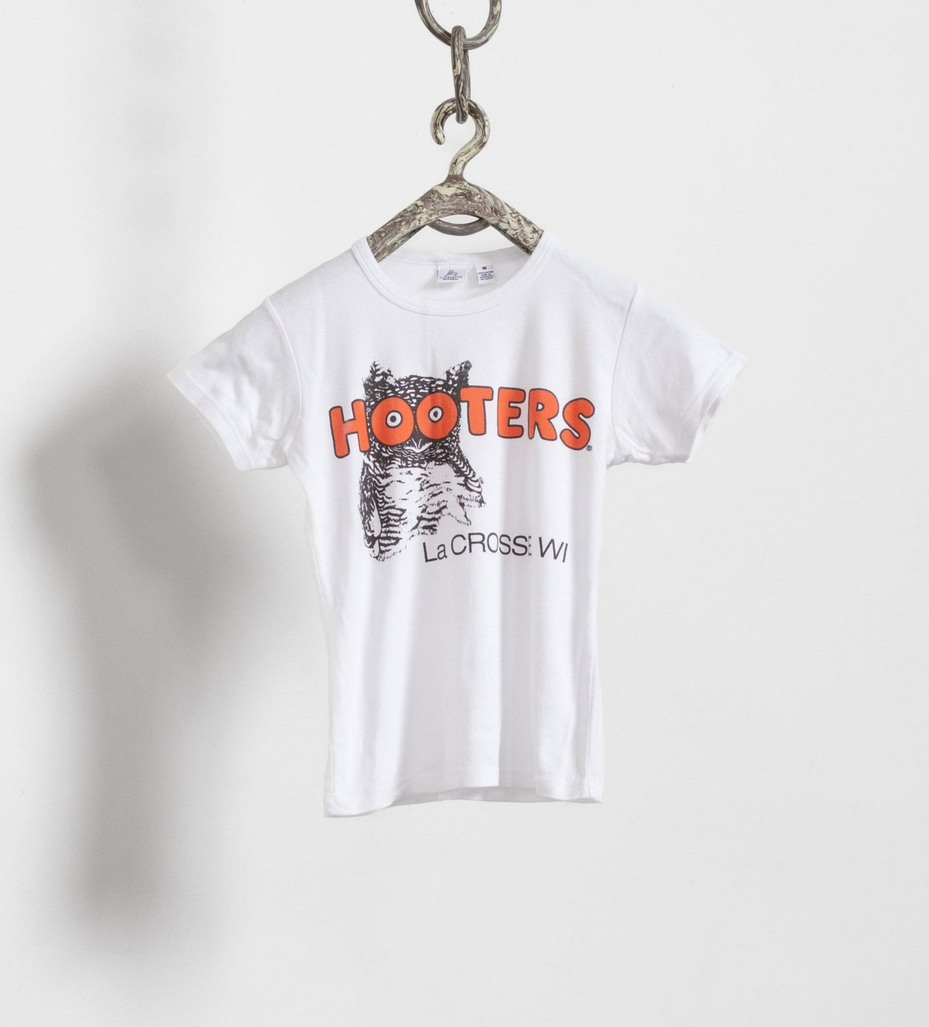 A white vintage Hooters t-shirt with orange and black text hanging from a chain against a plain background.