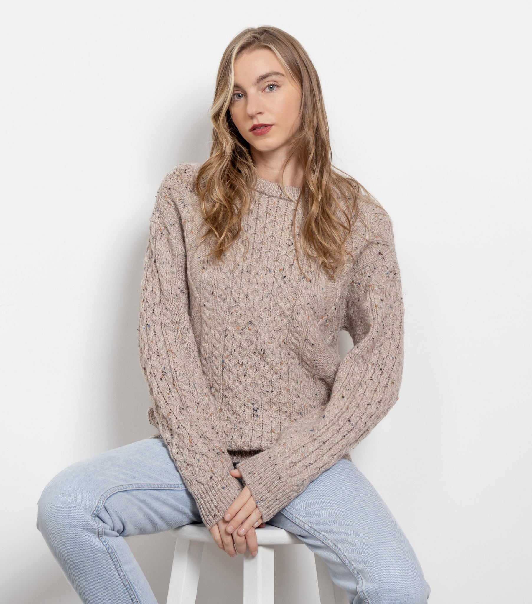 Woman wearing a beige cable knit sweater and light blue jeans sitting on a white stool against a plain background.