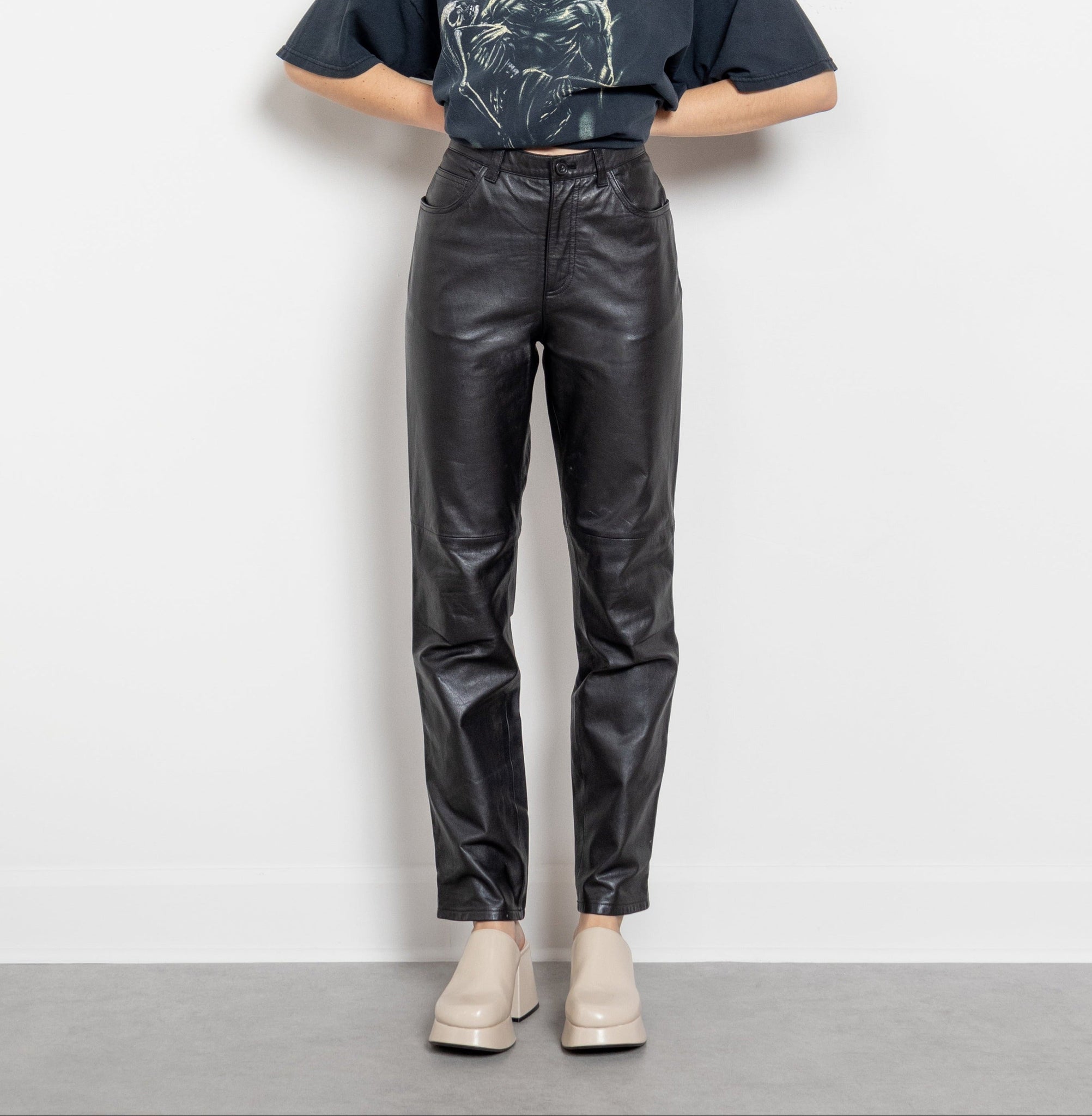 A person standing against a white background wearing high-waisted black leather pants with two front pockets and two back pockets, paired with a gray shirt and beige shoes.