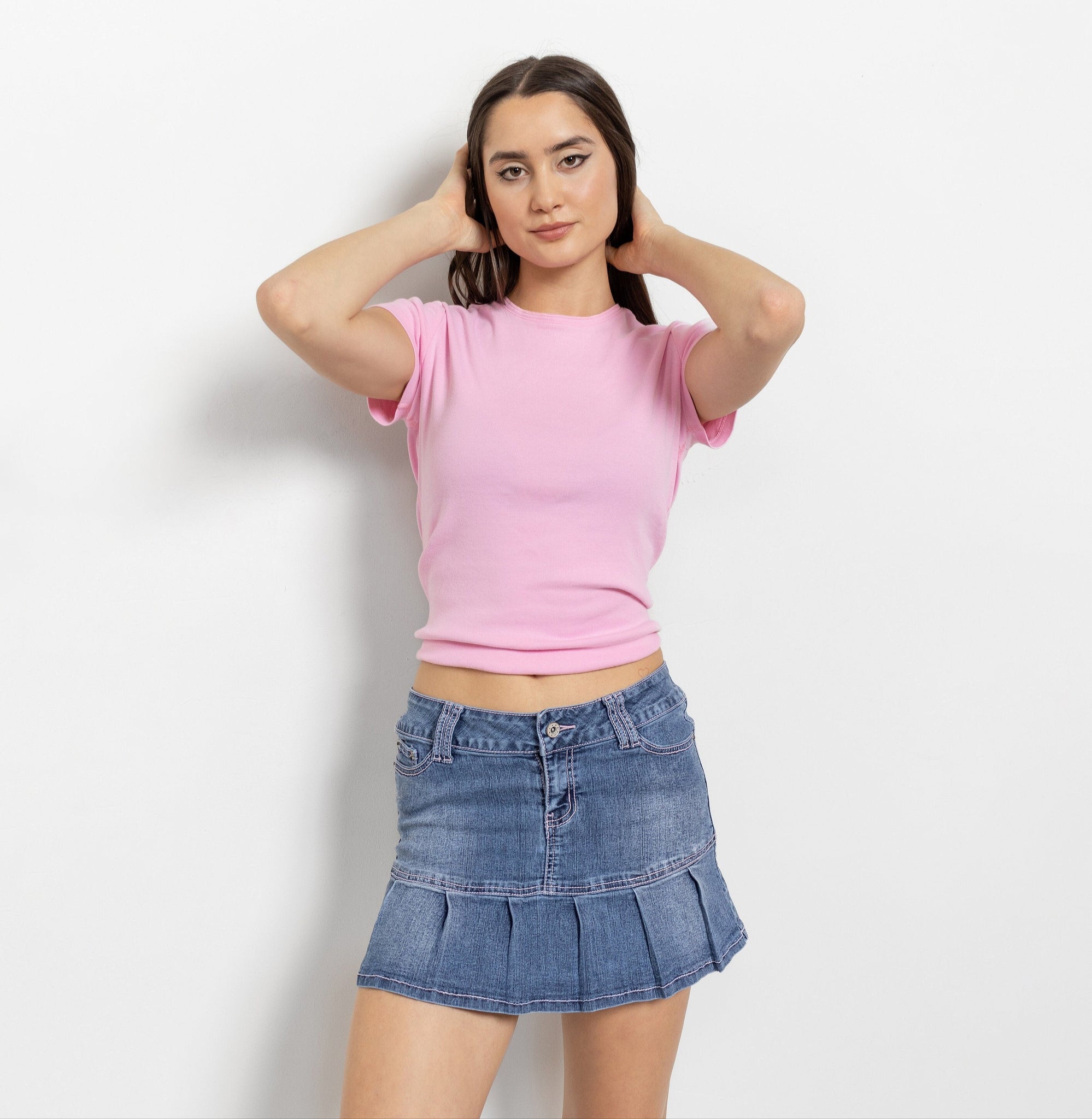 A person wearing a pink top and a blue denim mini skirt with pleats and a low-rise waist.
