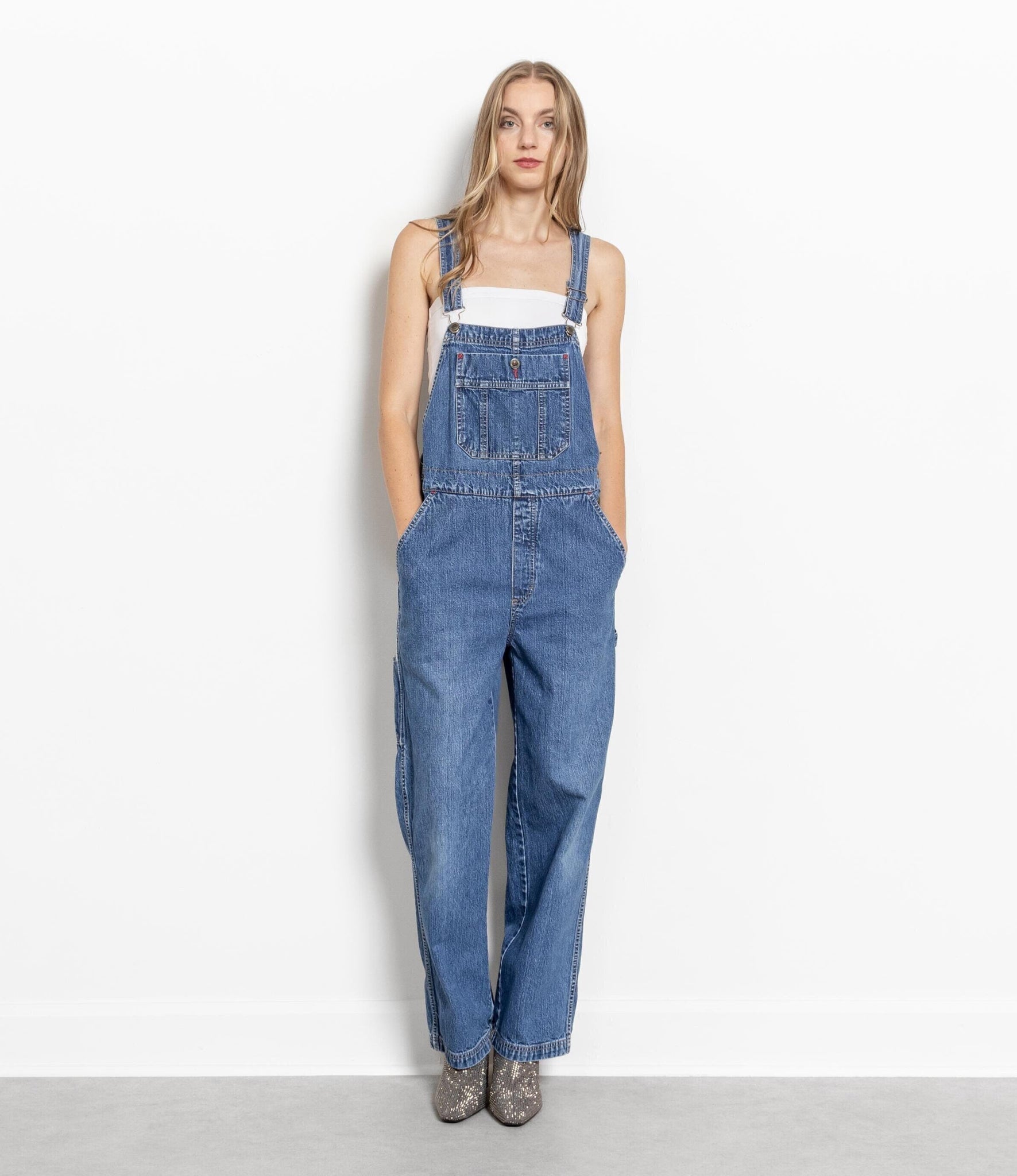 A female model wearing vintage baggy denim overalls with a loose fit, standing against a white background.