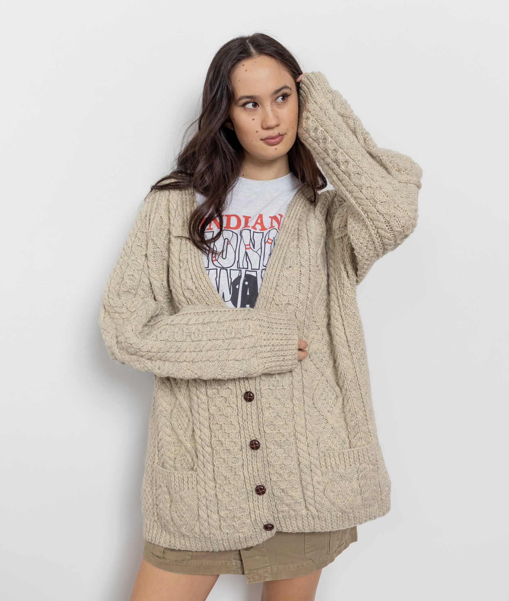 A woman wearing a beige cable knit Irish wool cardigan with a graphic t-shirt underneath and tan shorts. The cardigan is buttoned up the front and has two front pockets.