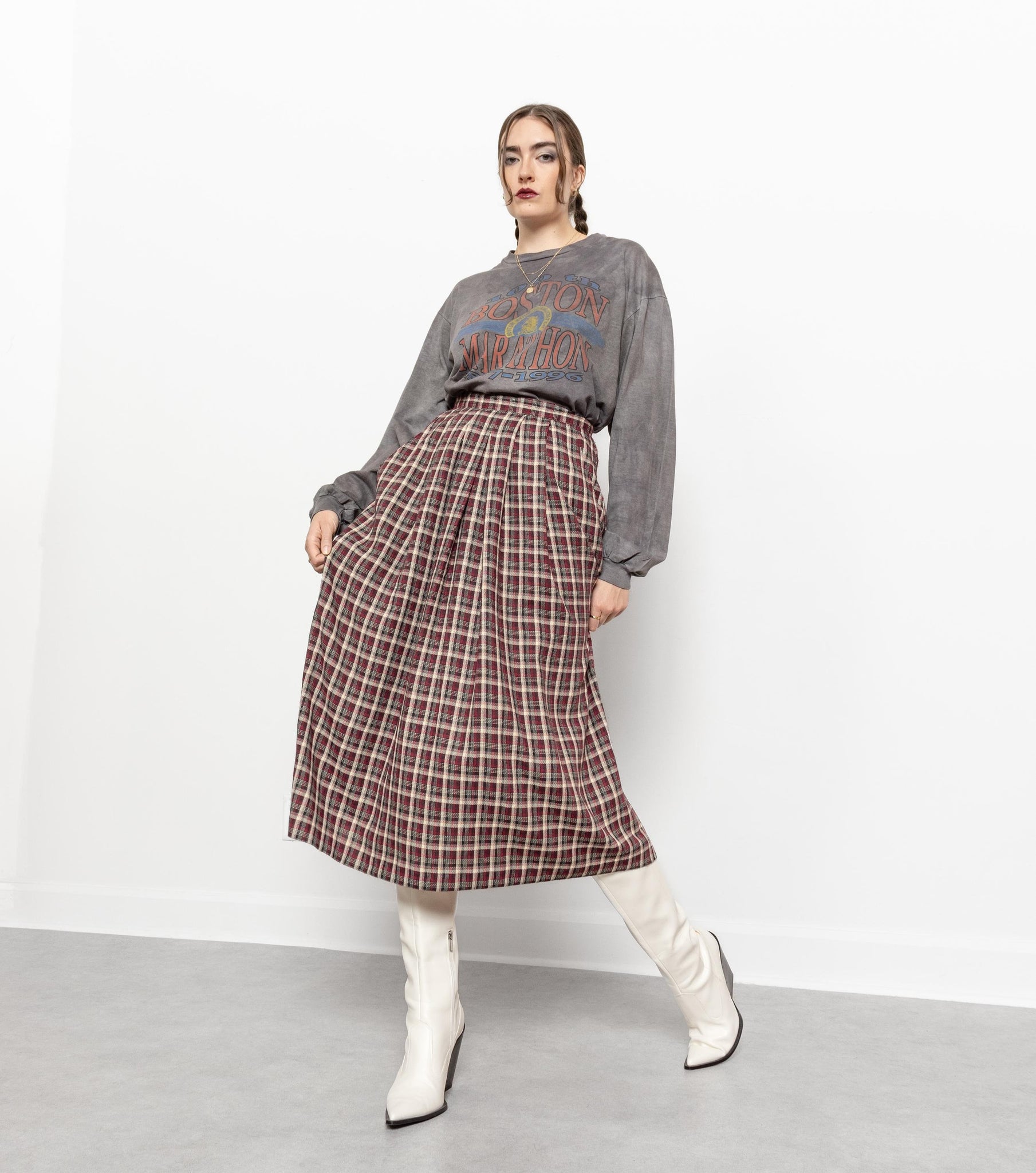 A person standing in a studio setting wearing a vintage Orvis plaid maxi skirt with a high waist and side pockets, paired with a grey sweater and white boots.