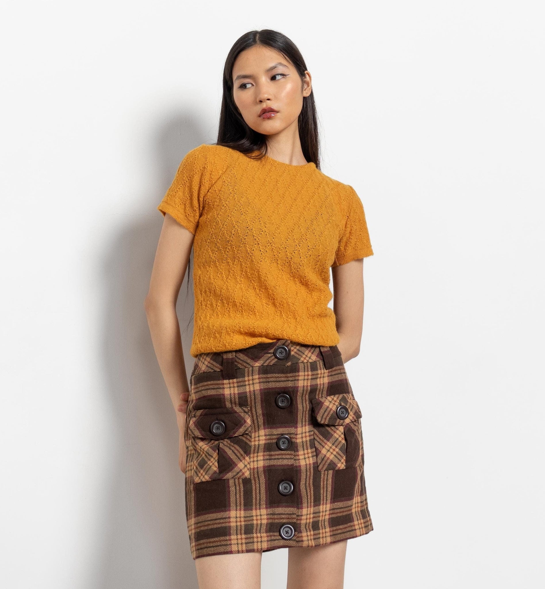 Woman wearing a mustard yellow top and plaid skirt against a white background
