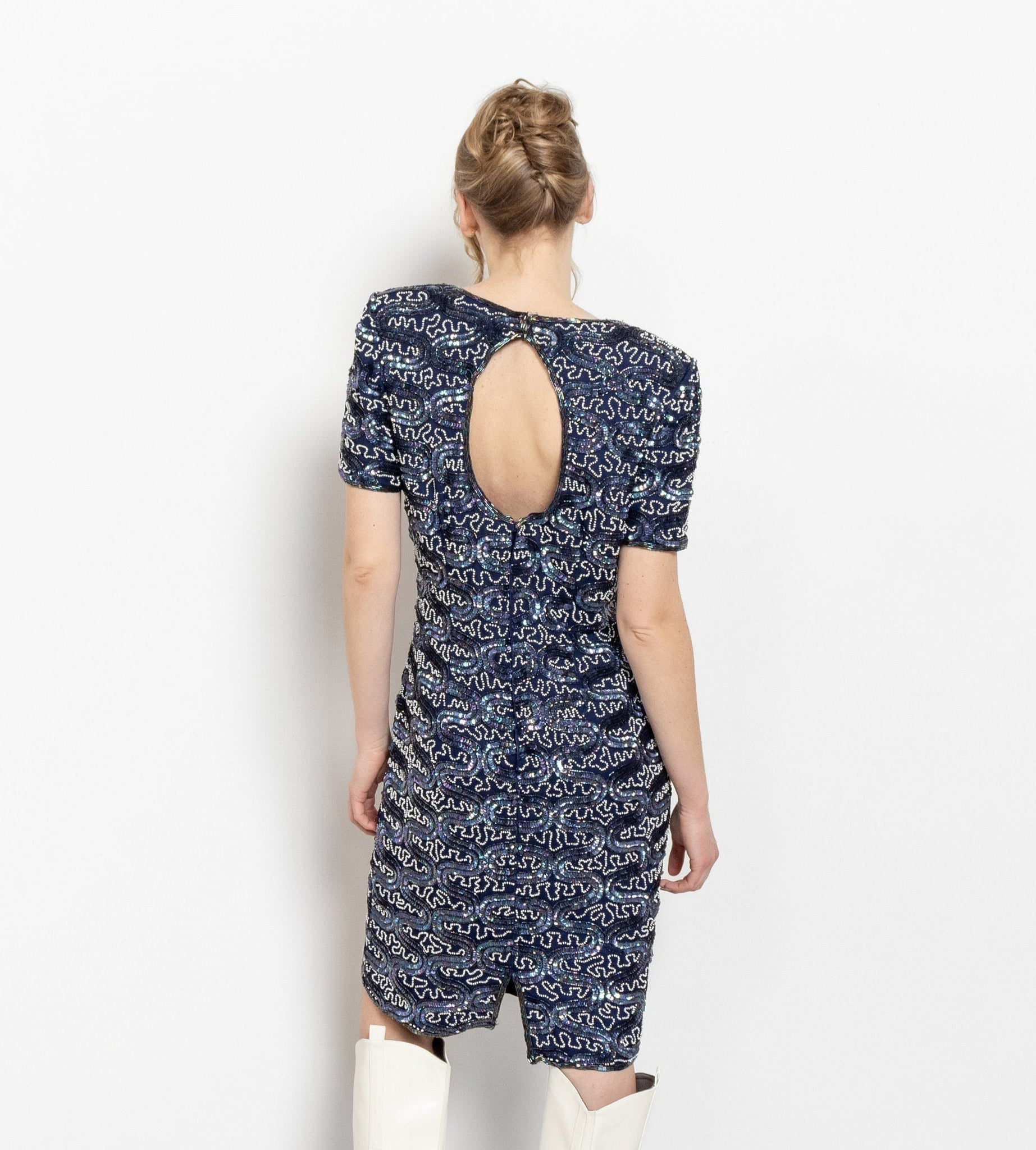 A woman viewed from the back wearing a vintage blue silk sequin dress with short sleeves and a mid-length design, featuring a keyhole back closure.