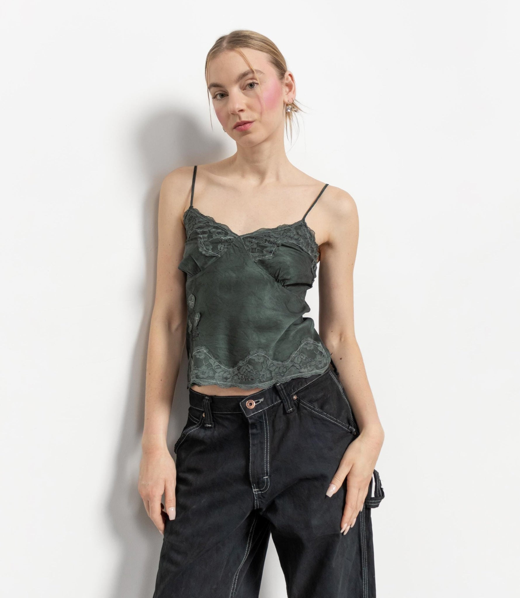 A woman modeling a dark green silk lace camisole with adjustable spaghetti straps and a flattering fit, paired with casual black jeans.