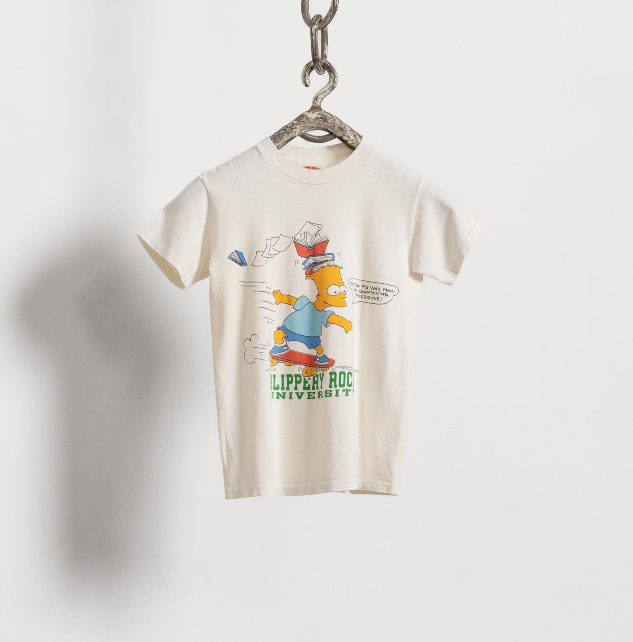 A vintage white Bart Simpson graphic t-shirt with a stack of books on his head and a text bubble, hanging on a hanger against a white background.