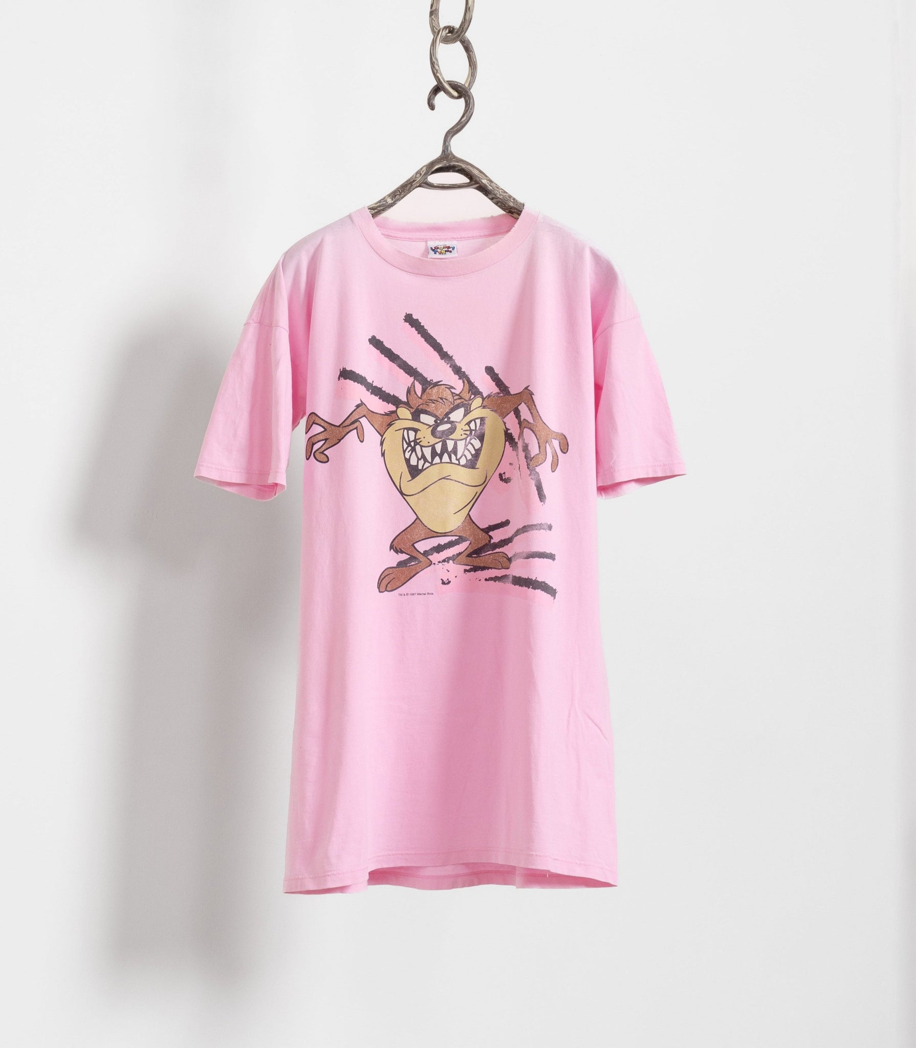 A pink t-shirt with a vintage longline cut and a graphic of a Tasmanian devil character on the front, displayed on a hanger against a white wall.