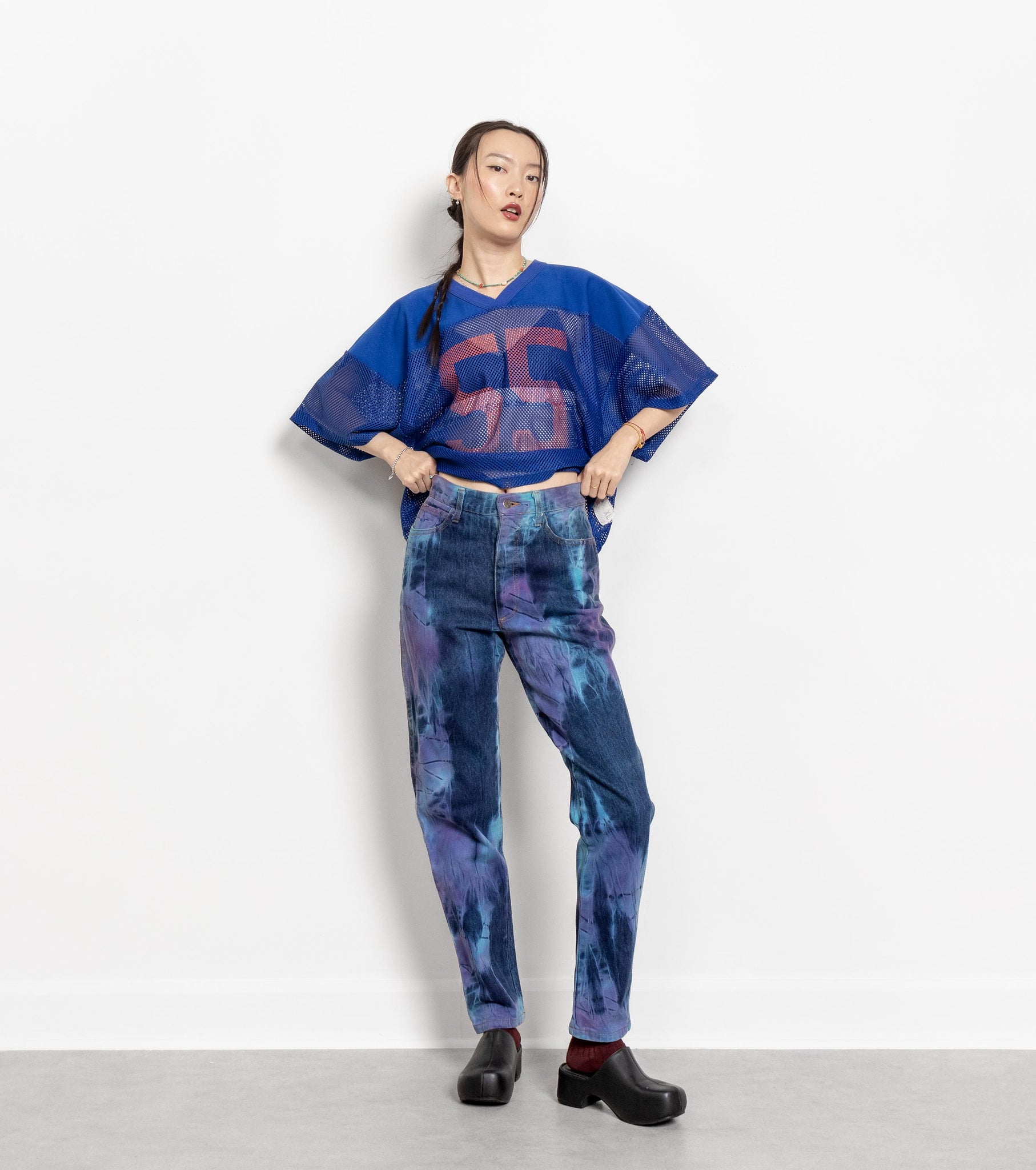 A person standing against a white background, wearing a tie-dye blue and purple shirt with high-waisted straight-leg jeans.