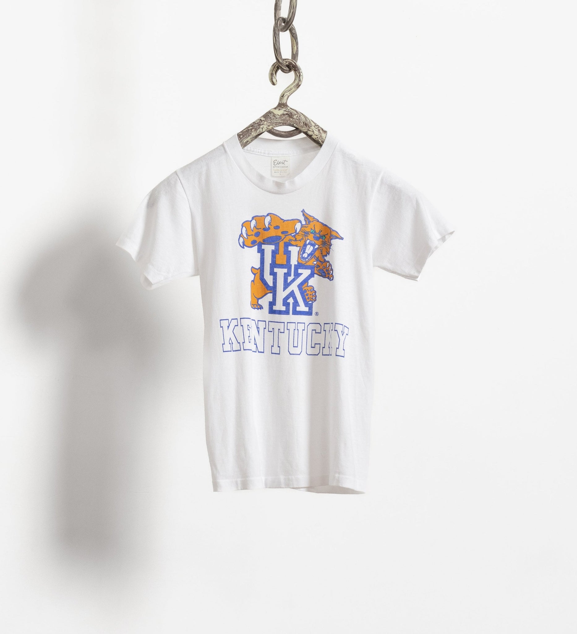A white University of Kentucky collegiate t-shirt with a graphic design, hanging from a chain against a white background.