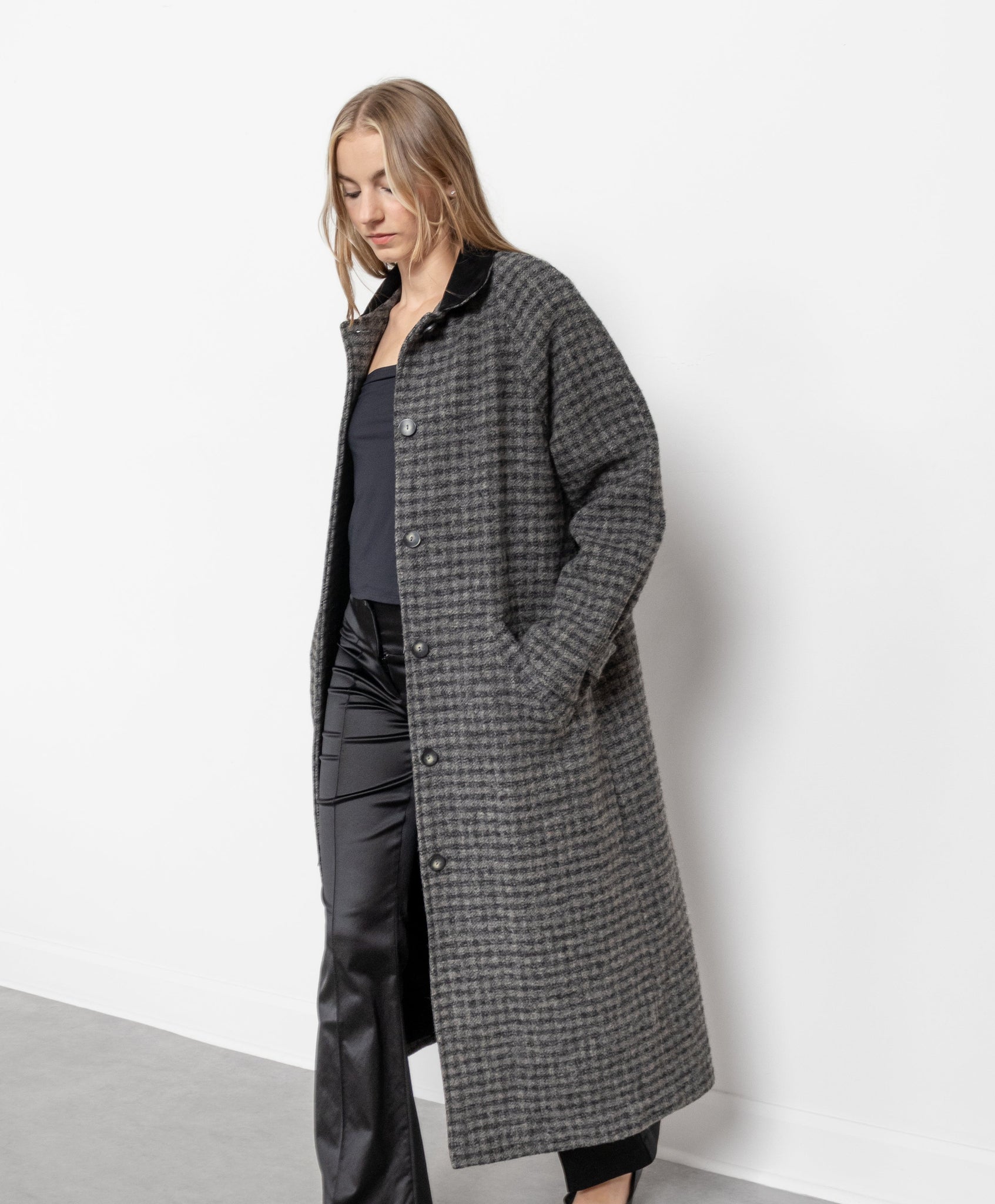 A person standing in a studio setting wearing a vintage 1990's wool blend gingham trench coat in shades of grey with a contrasting dark grey suede collar, buttons down the front, and side pockets.
