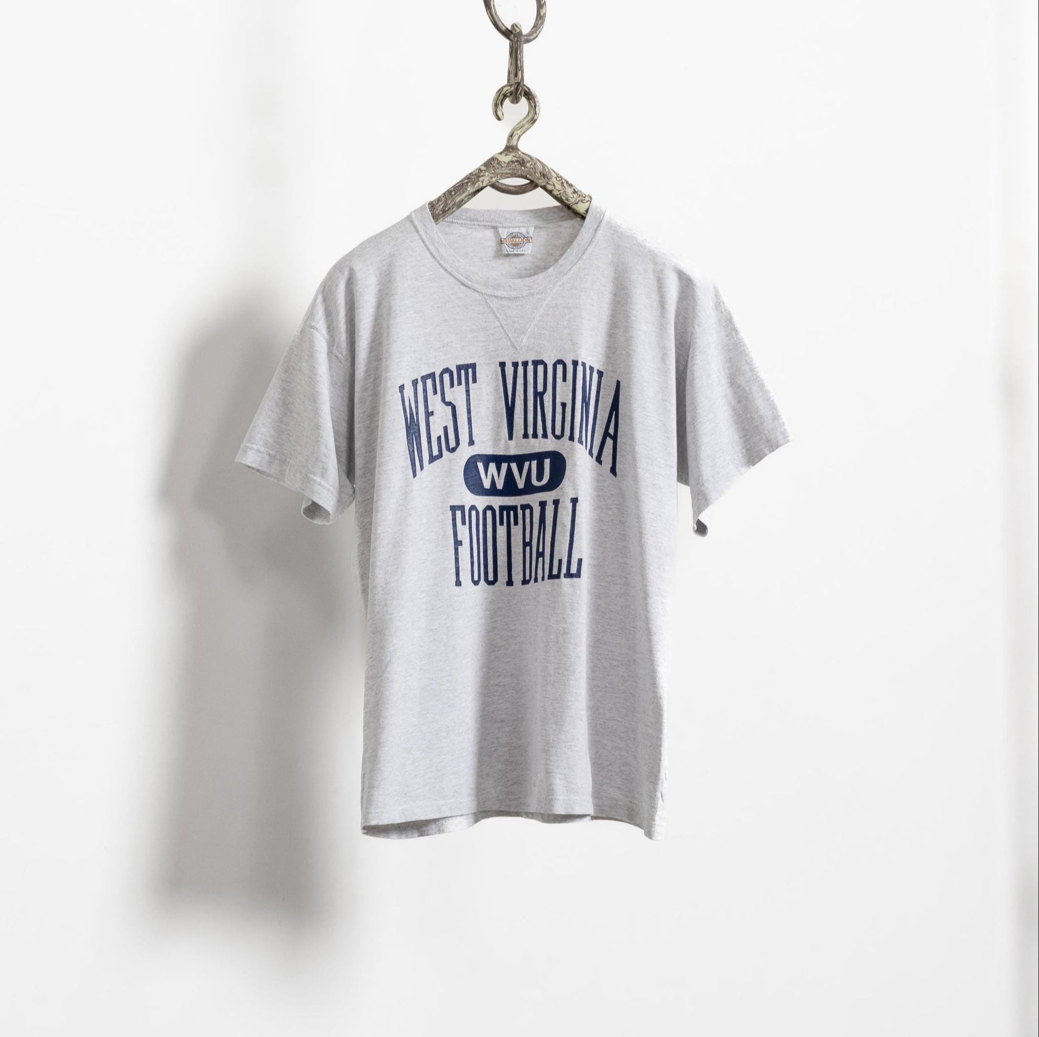A vintage short sleeve gray t-shirt with 'West Virginia Football' printed in navy blue on the front, displayed on a hanger against a white background.