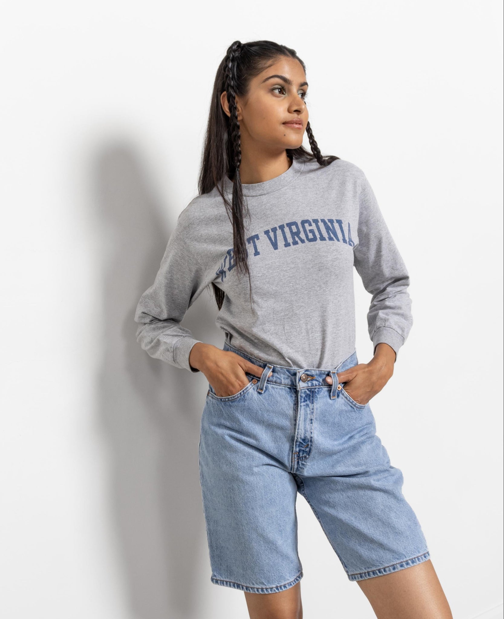 A person wearing a gray long sleeve crew neck t-shirt with 'WEST VIRGINIA' printed in blue letters on the front, paired with blue denim shorts.