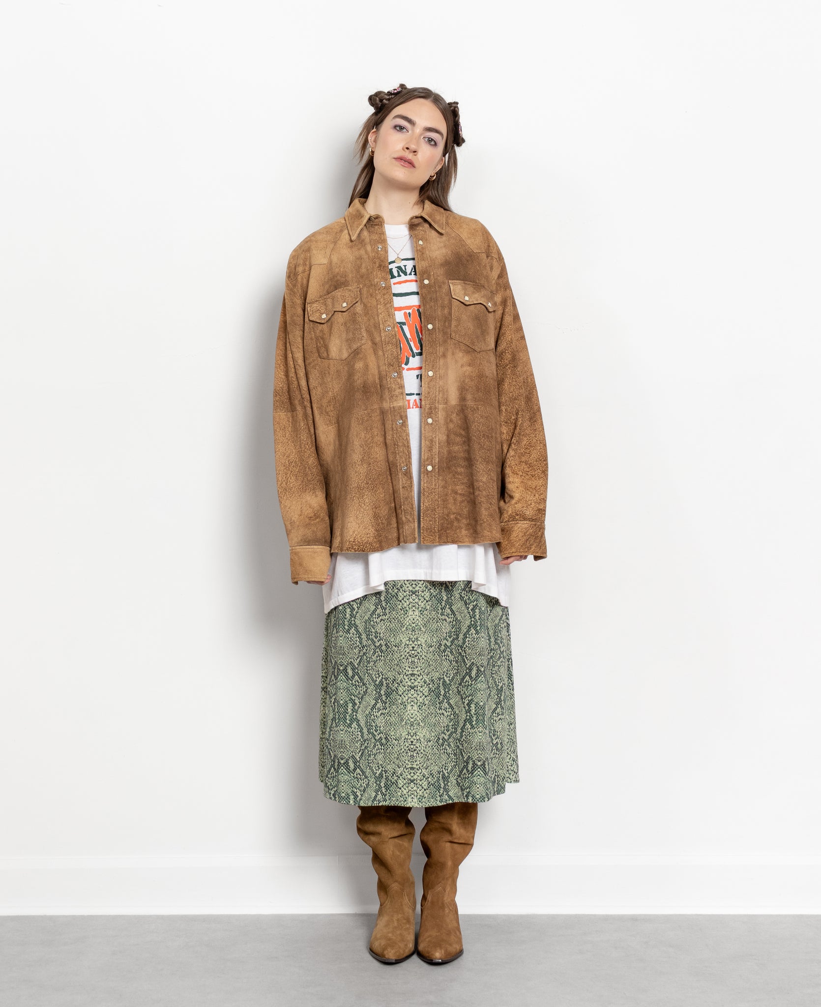 Person wearing a brown suede jacket, white shirt, patterned skirt, and brown boots on a white background