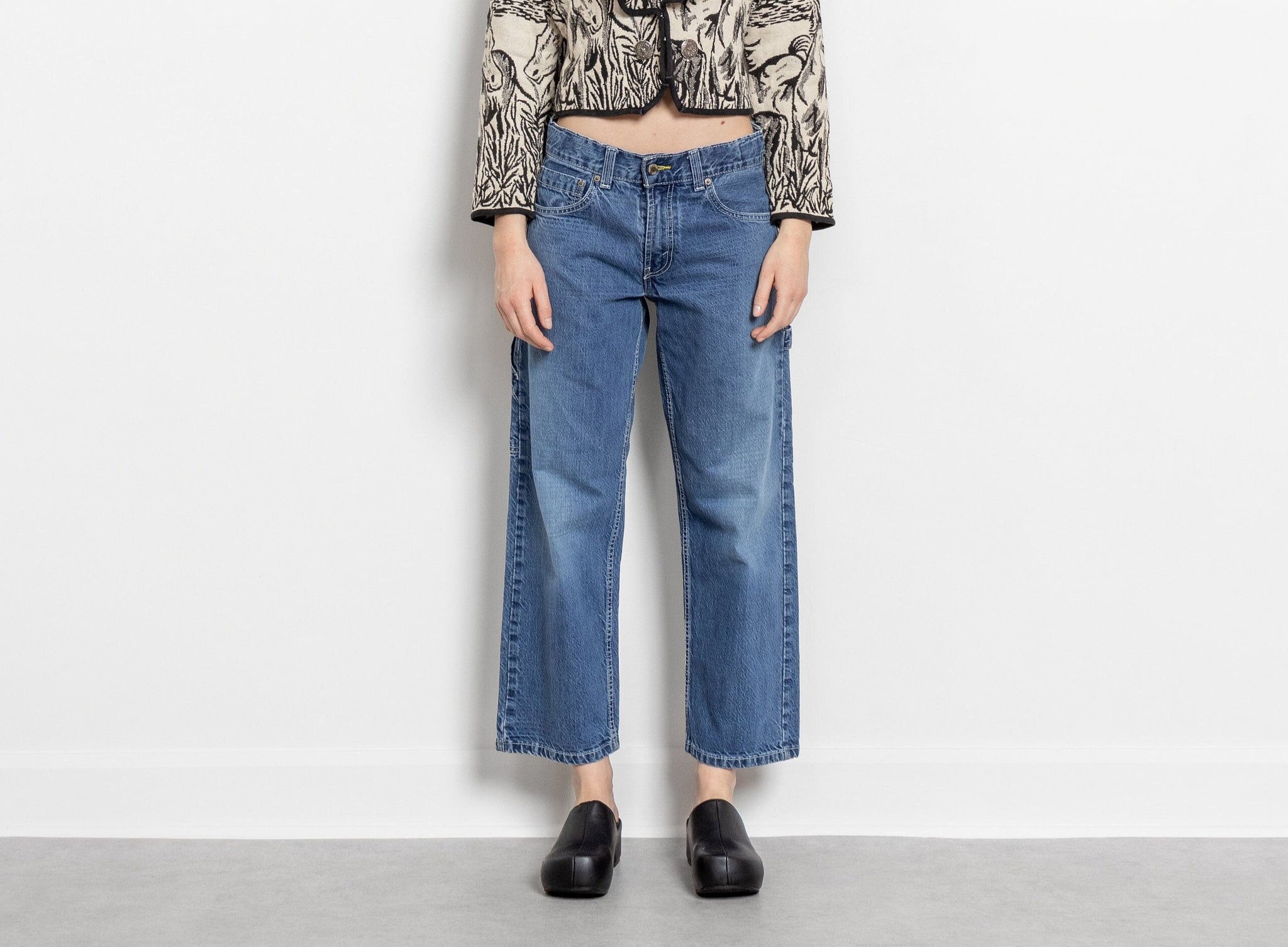 A person standing against a white background, wearing medium wash, low/mid rise Levi's jeans with a wider relaxed leg, paired with a patterned top and black shoes.