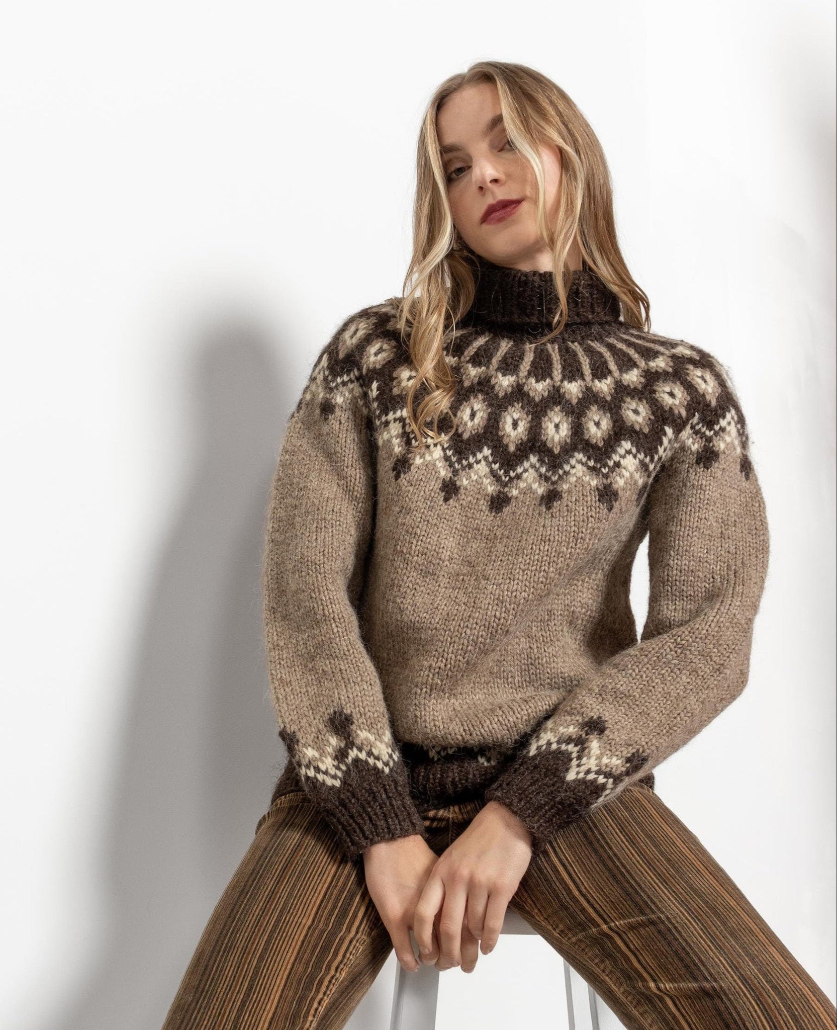 A person seated on a stool wearing a beige and brown striped sweater with a fair isle knit pattern, paired with brown trousers.