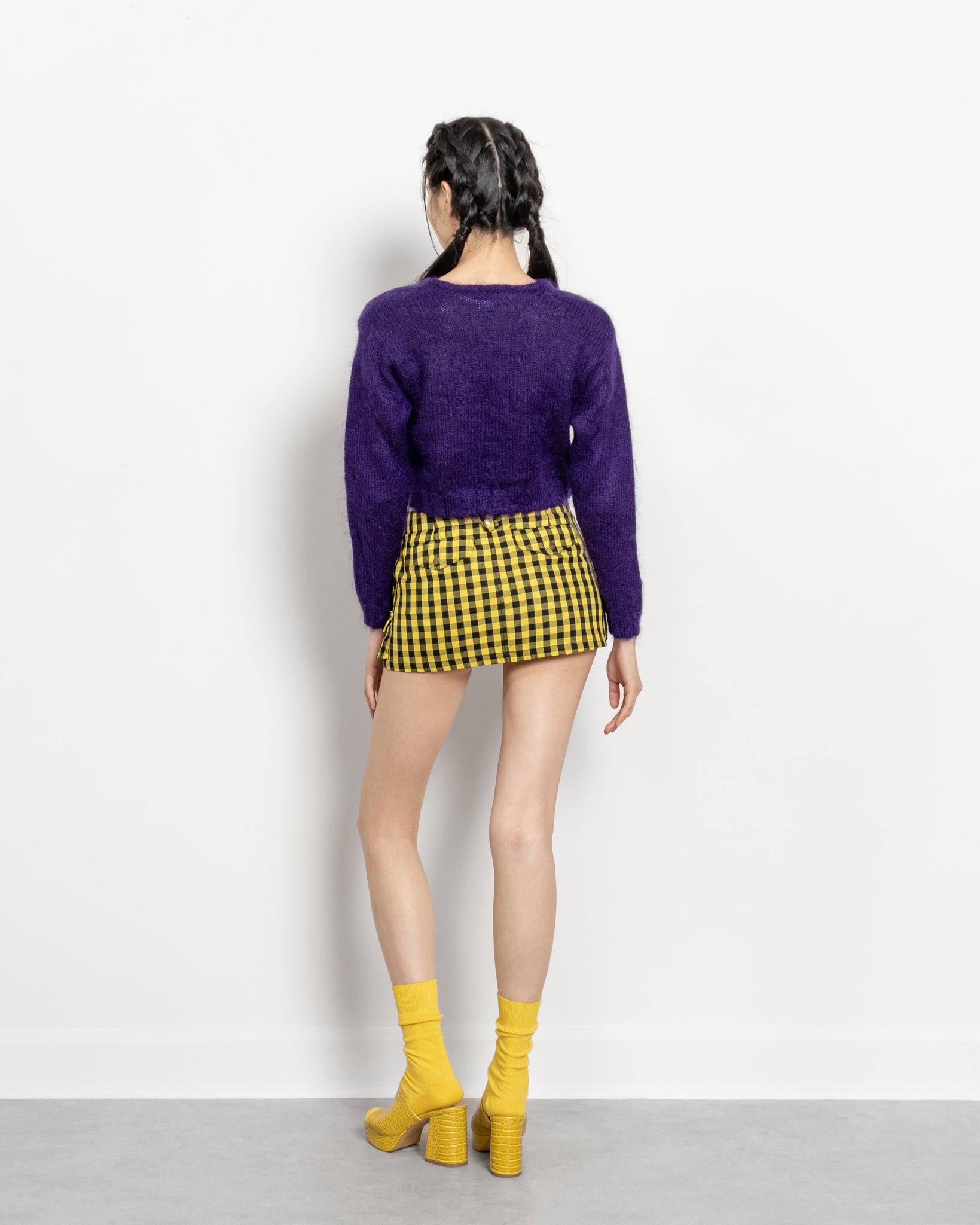 Person wearing a purple sweater and yellow checkered skirt with yellow shoes on a white background