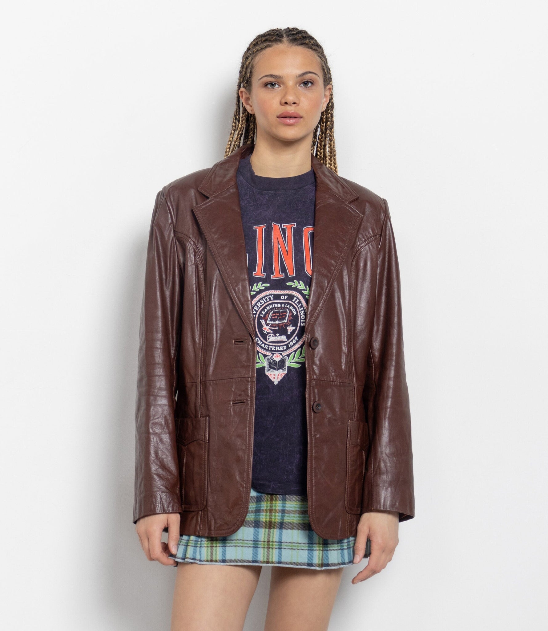 Person wearing a brown leather jacket over a graphic t-shirt and plaid skirt on a white background