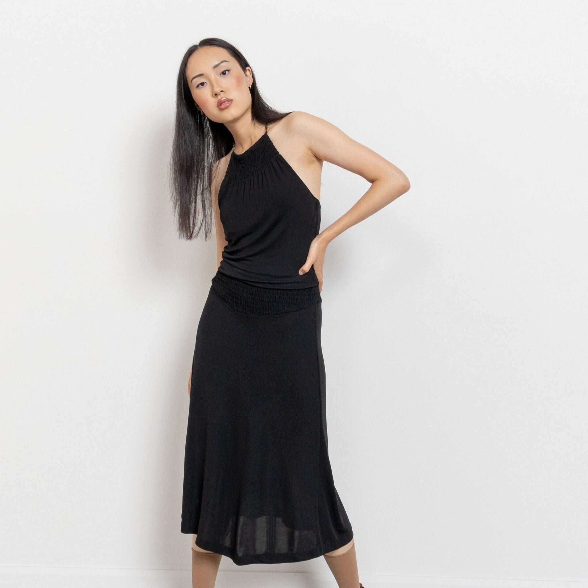 A person posing in a vintage 90's black stretch halter dress with a gathered/smocked elastic waist and a flared skirt, paired with ankle strap sandals.