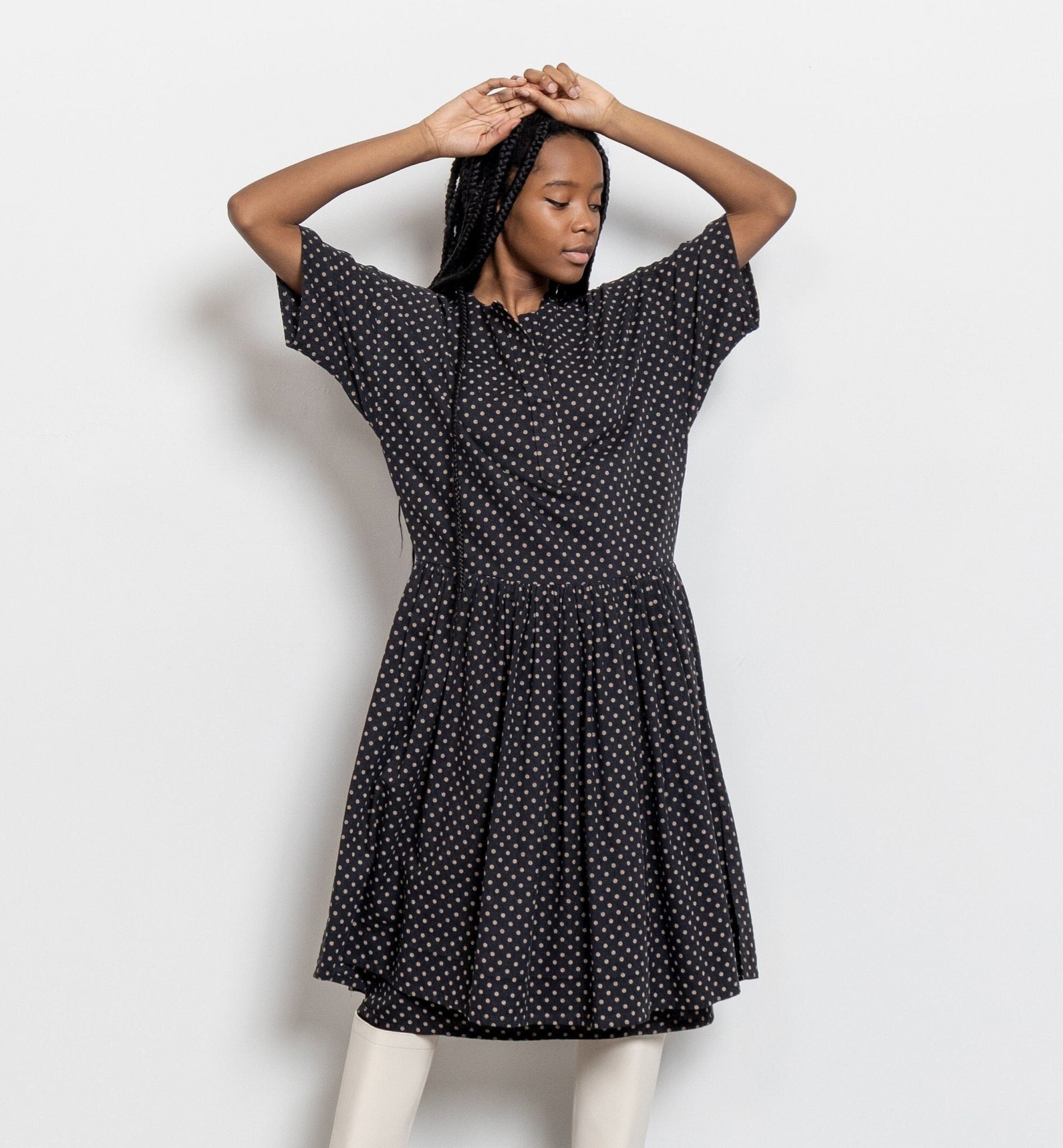 A woman standing in a studio setting, wearing a vintage polka dot cotton midi dress with a gathered skirt and half-length sleeves. She is posing with one hand raised to her head and is wearing white boots.