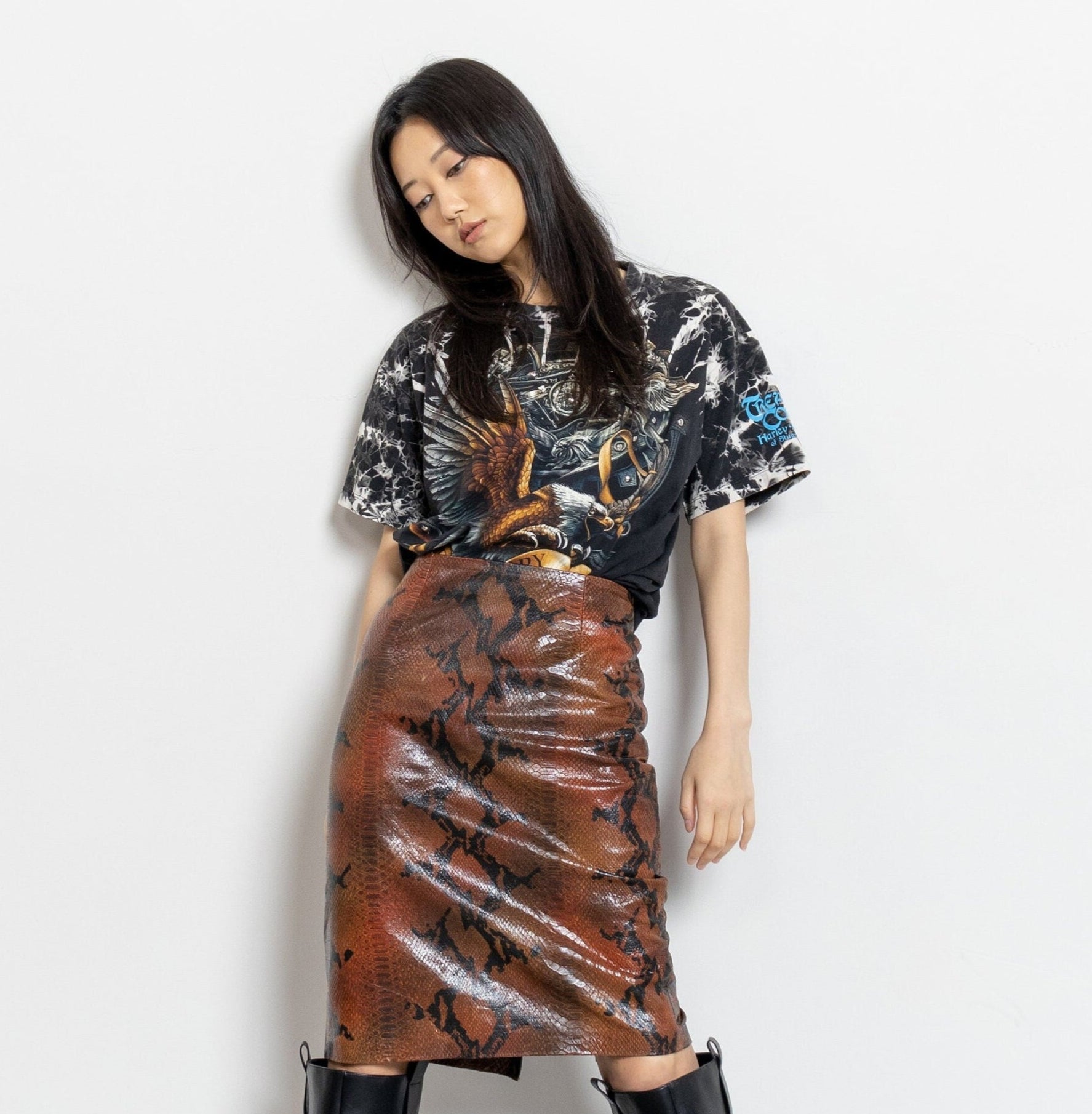 A person standing against a white background, wearing a high-rise leather midi skirt with a snake print design, paired with a multicolored shirt and black boots.