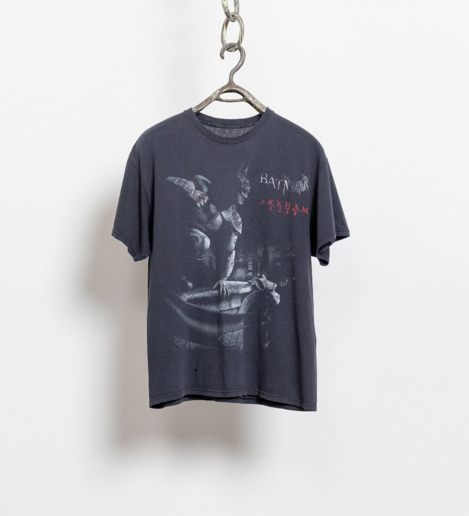 A gray Batman Arkham City graphic t-shirt hanging against a white background, showcasing natural distressed details and a variety of holes in the fabric.