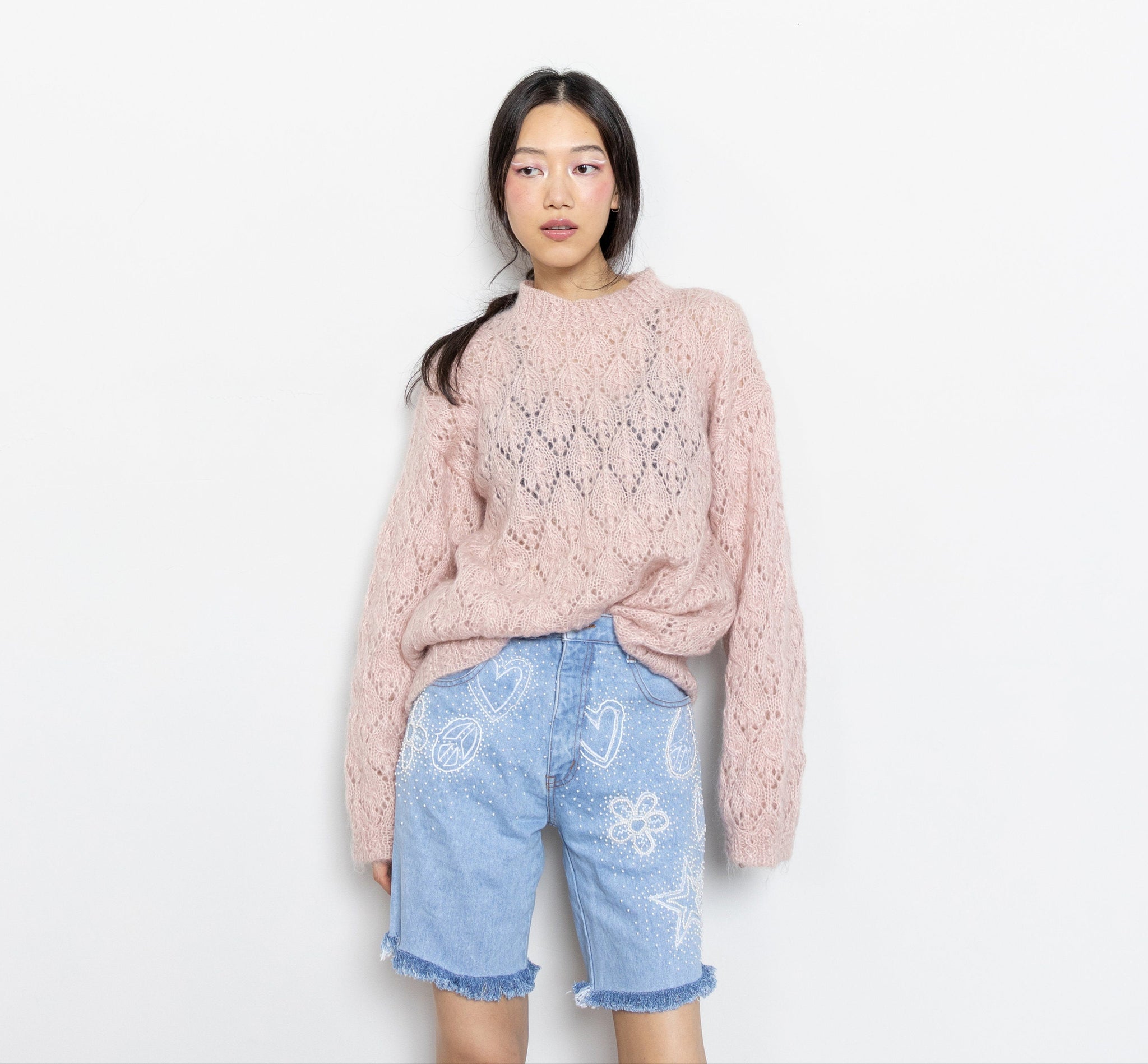 A person wearing a pink knit sweater and blue denim longline shorts with beaded details.