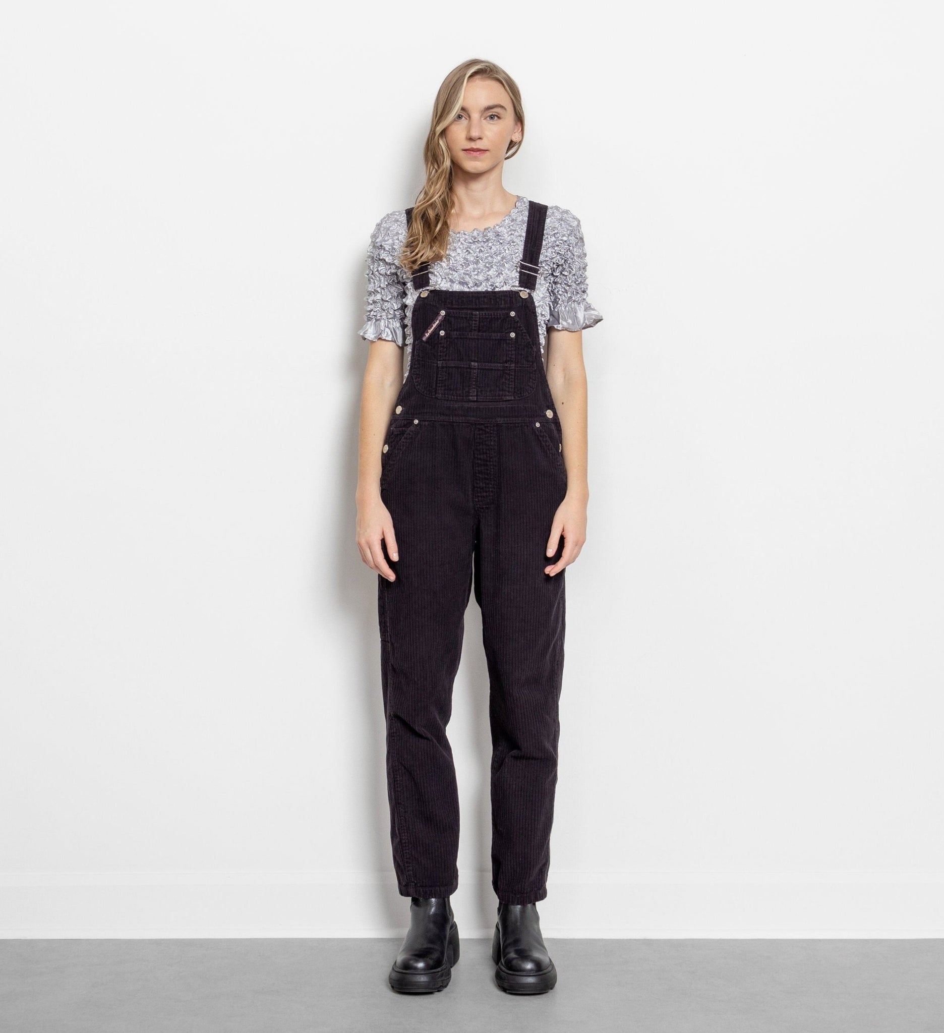 A person wearing vintage 90's black corduroy overalls with multiple pockets and adjustable straps, paired with black boots.