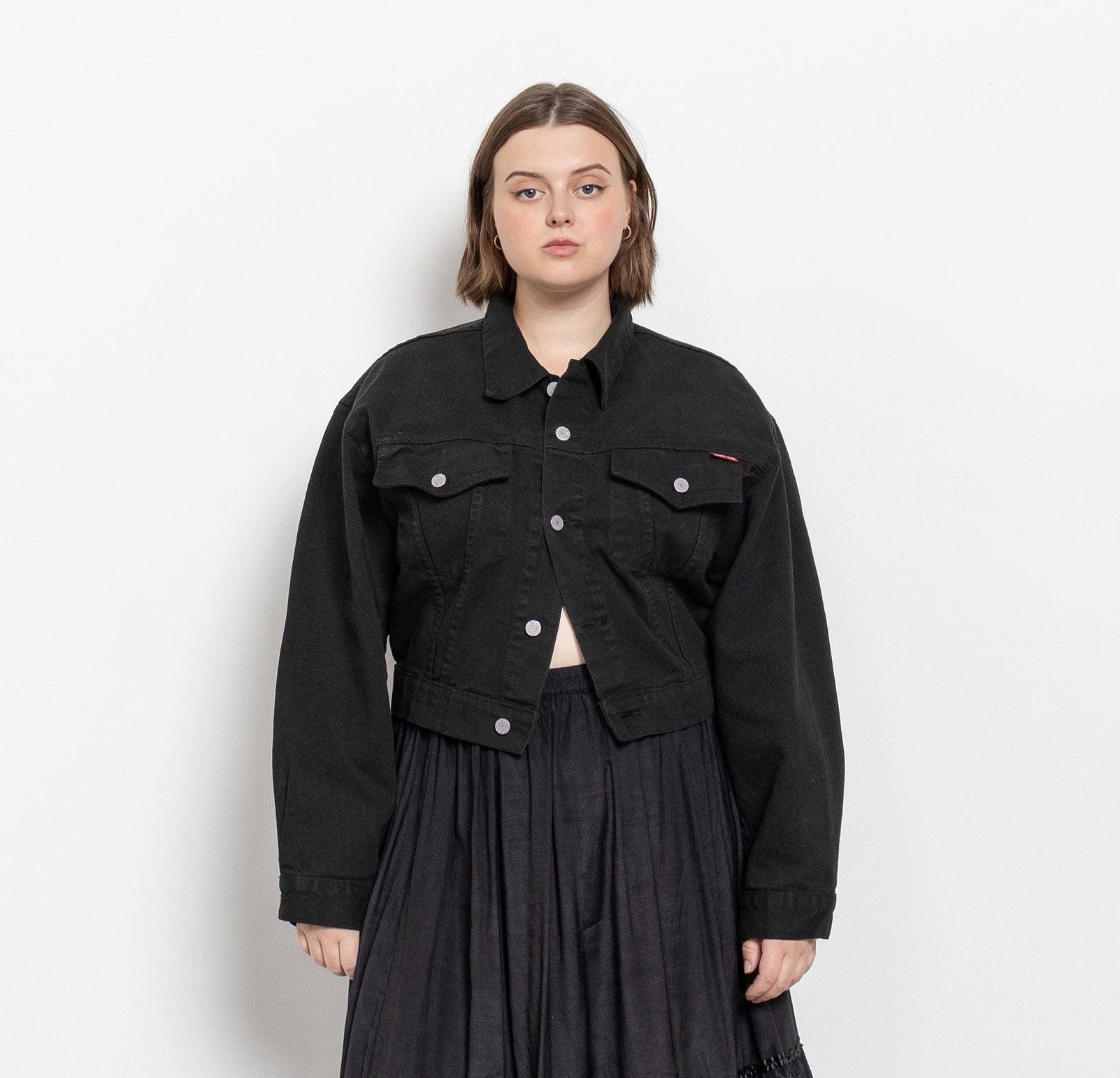 A person wearing a vintage 90's cropped black denim jacket with buttons up the front and breast pockets, paired with a dark skirt.