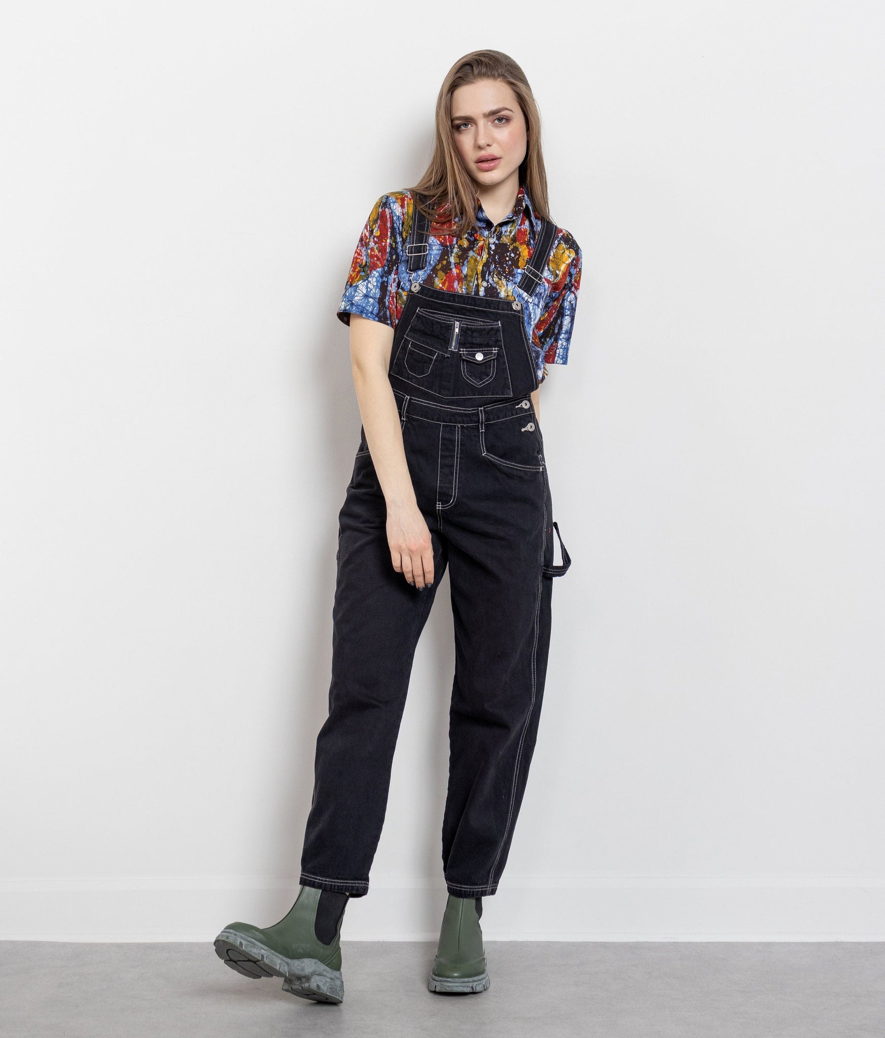 Person wearing black denim overalls and a colorful shirt on a white background