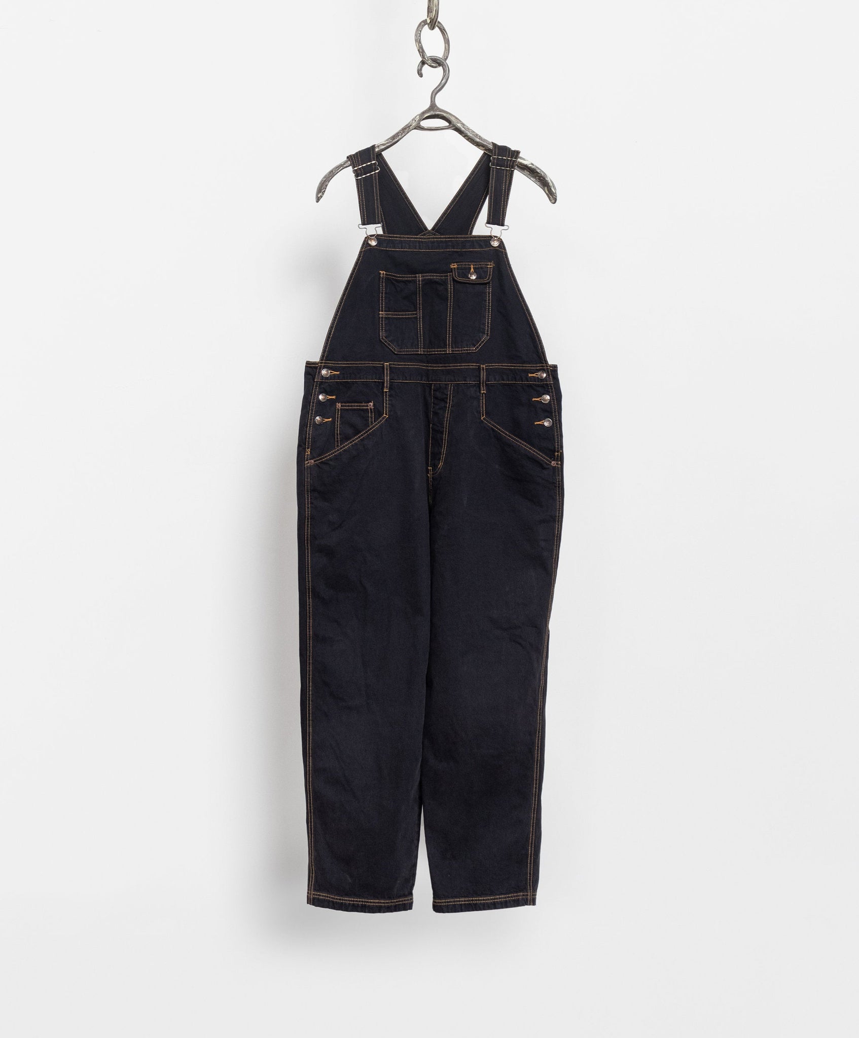 A pair of vintage 90's black denim overalls with contrasting yellow stitching, hanging on a hook against a white background.