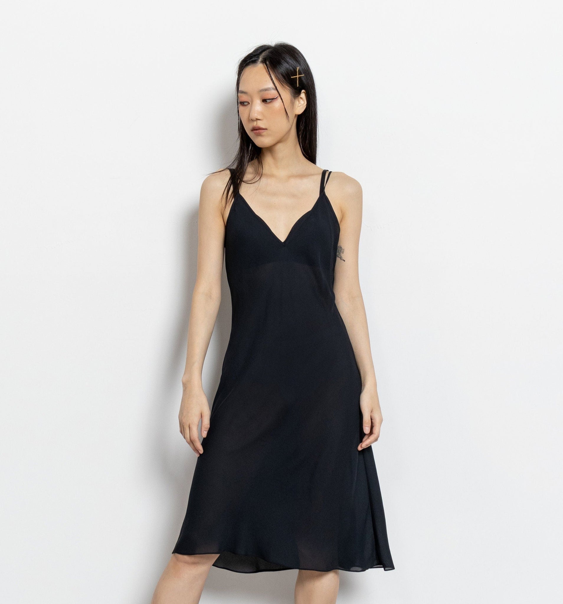 A model wearing a sheer black mini dress with spaghetti straps and a plunging neckline, standing against a white background.