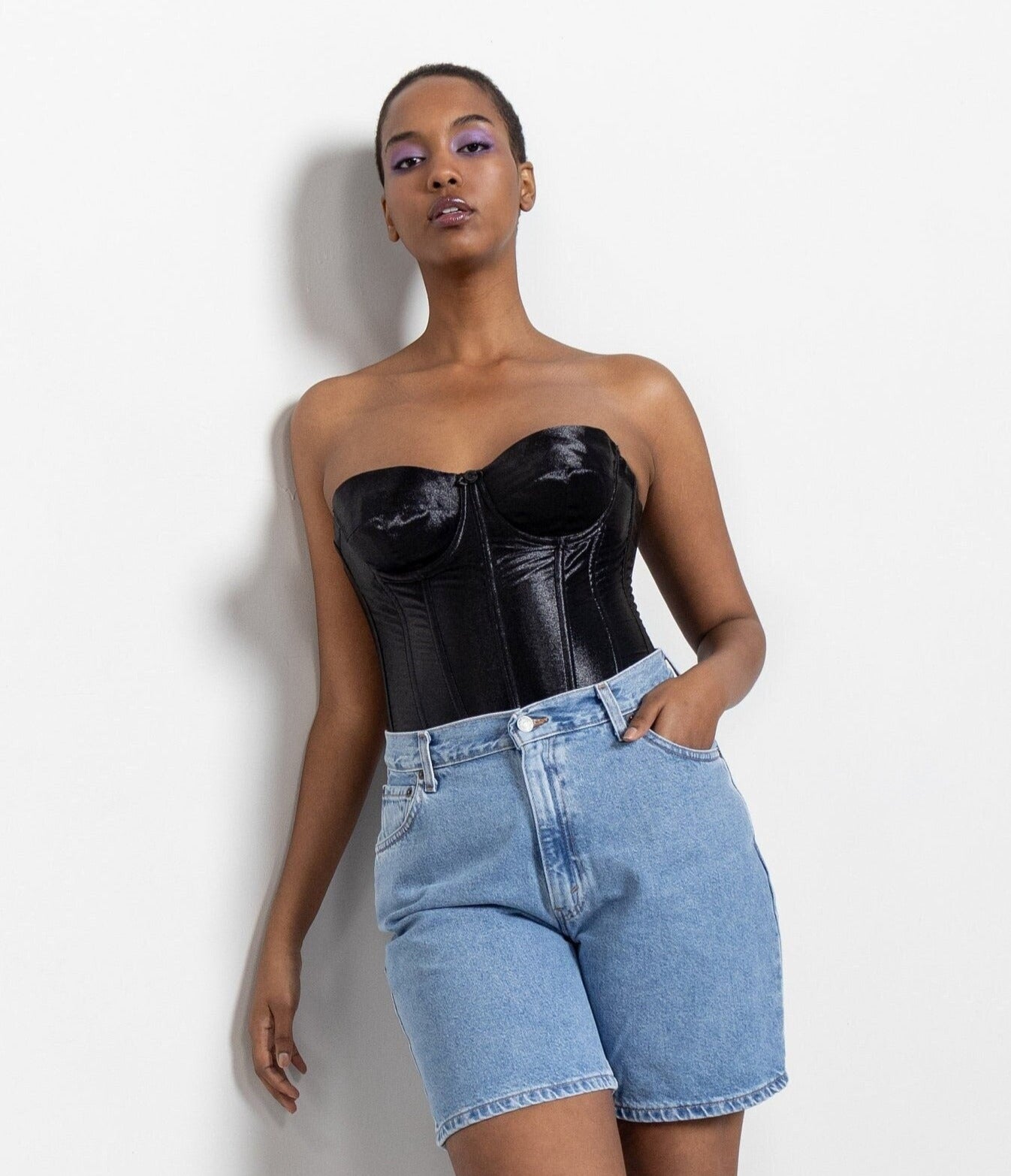 A model wearing a black satin corset top with wired padded cups and a sheer mesh back, paired with blue denim shorts.