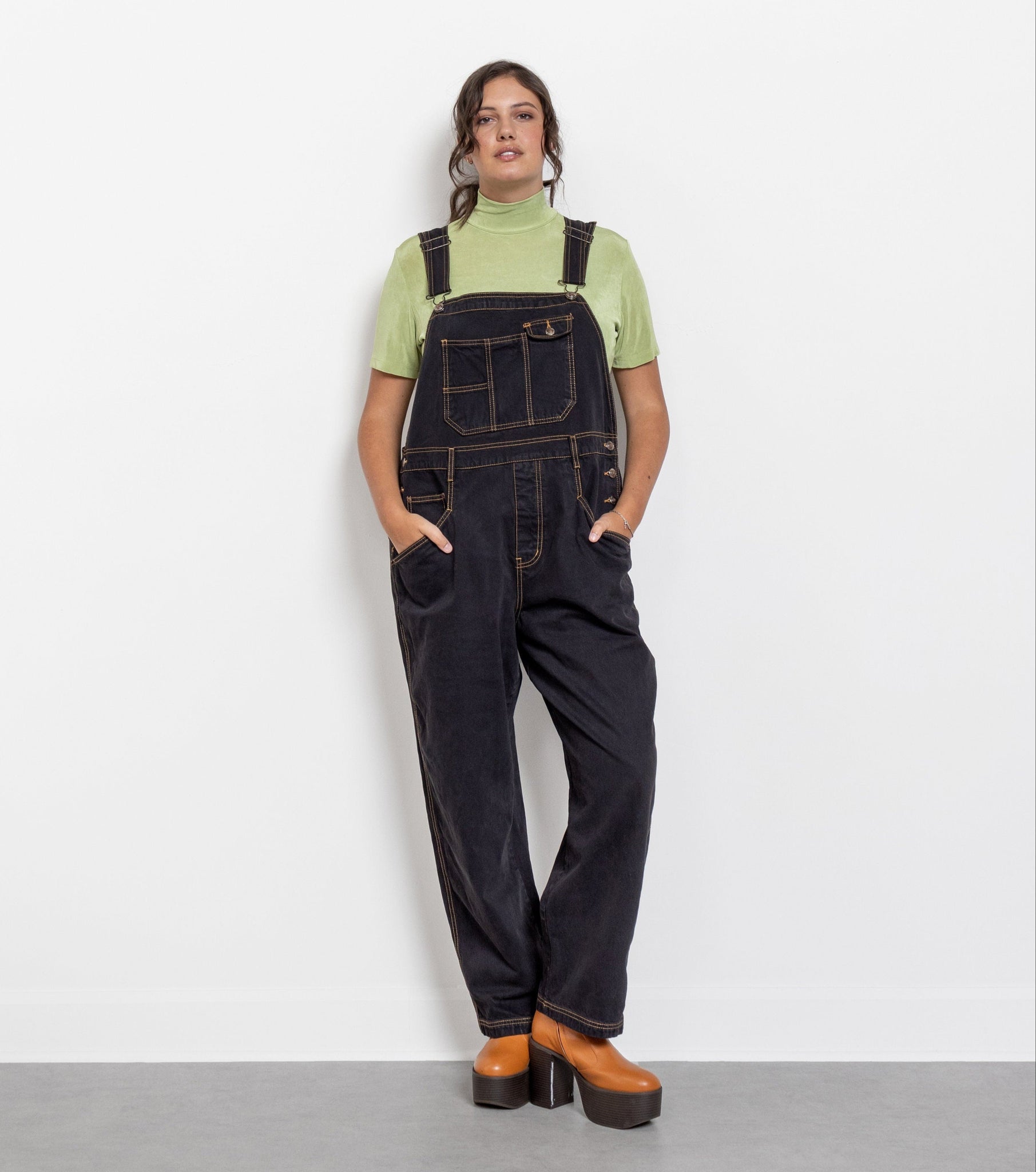 Person wearing black denim overalls and green shirt on a white background