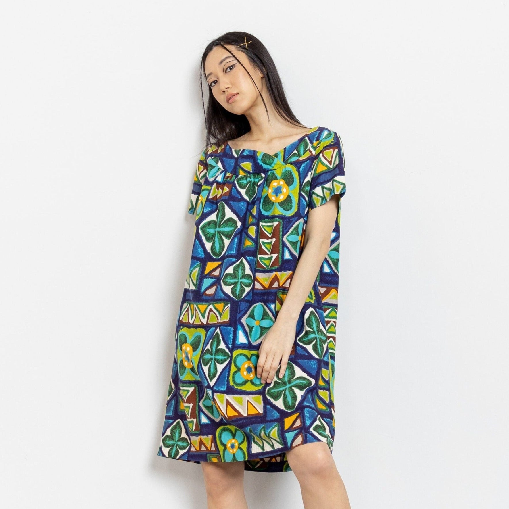 A woman modeling a vintage woven cotton tunic midi dress with a colorful abstract pattern in hues of green, blue, and yellow, featuring short sleeves and a pleated babydoll bust.
