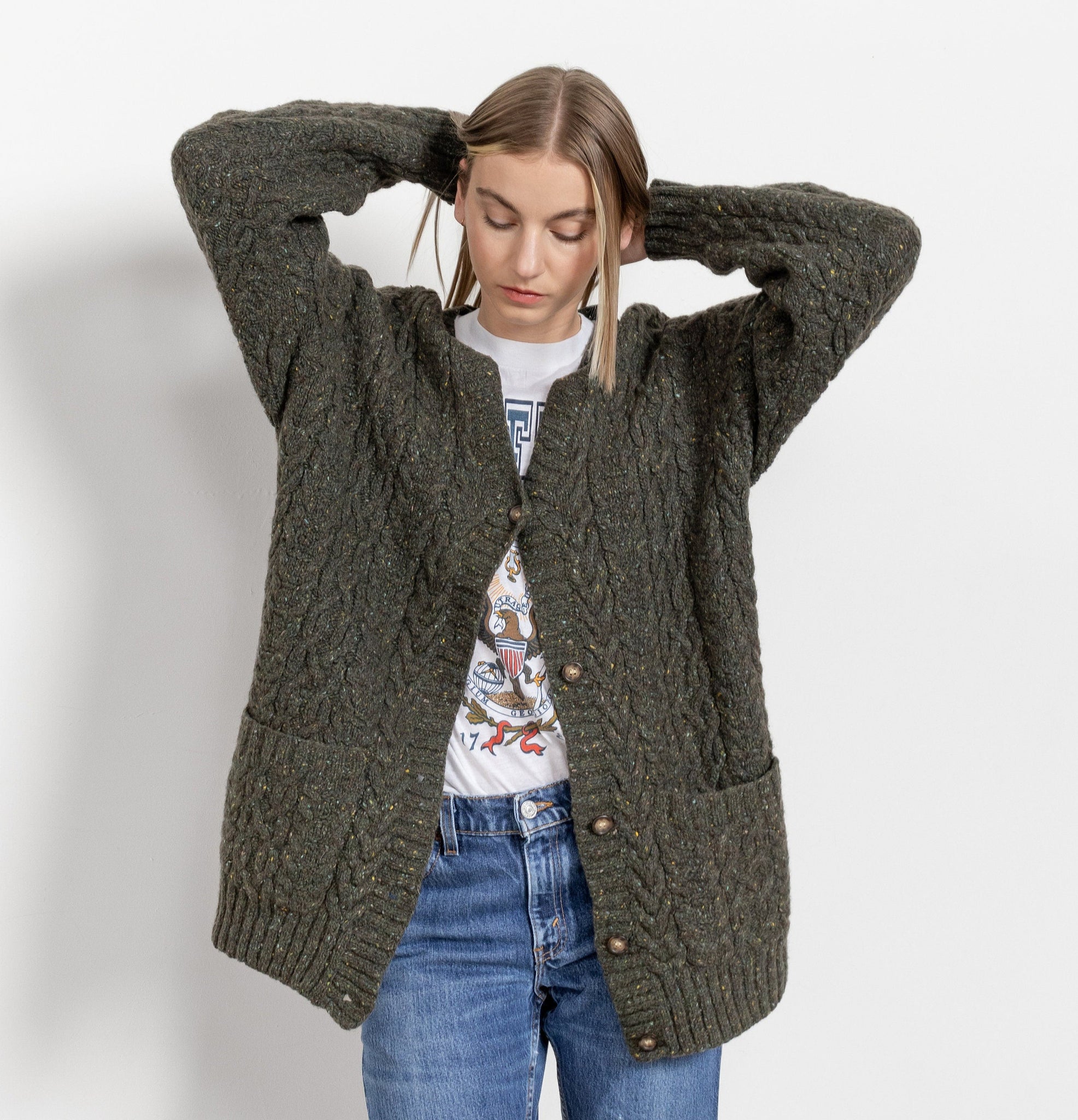 A woman wearing a vintage green cable knit traditional cardigan with a round neckline and front pockets over a white shirt and blue jeans.