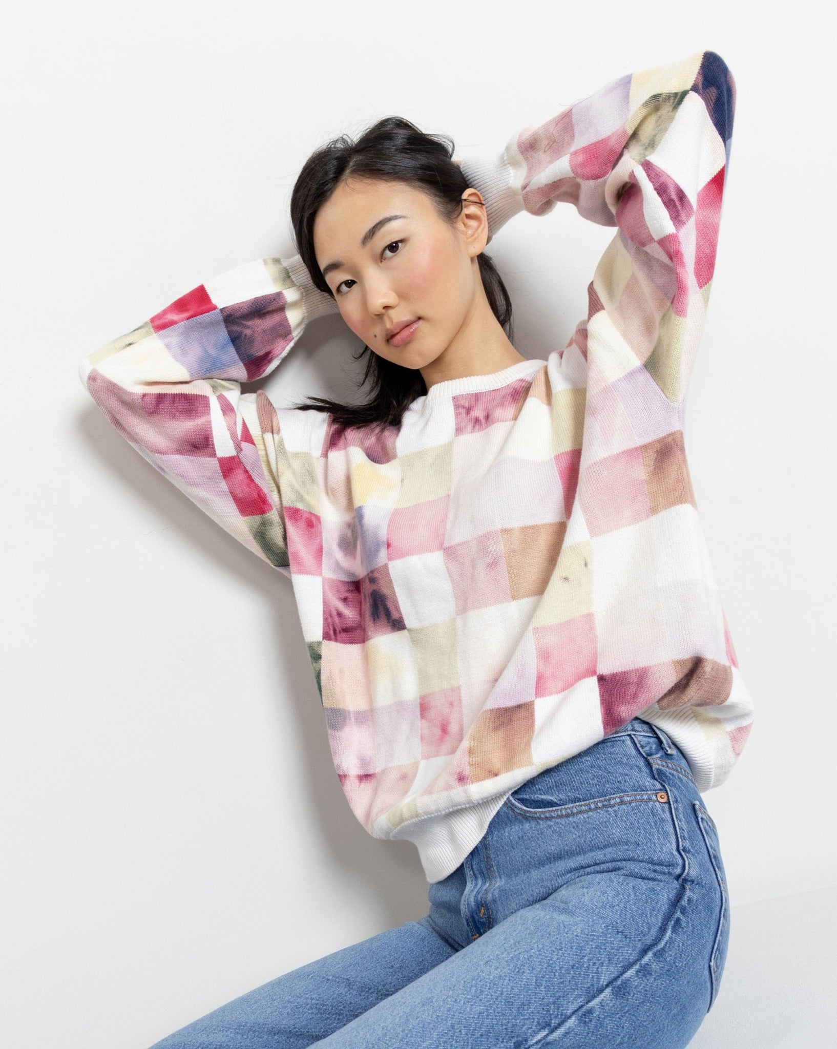 A woman sitting and posing with a pastel toned plaid 100% cotton knit sweater in pink and white checkers.