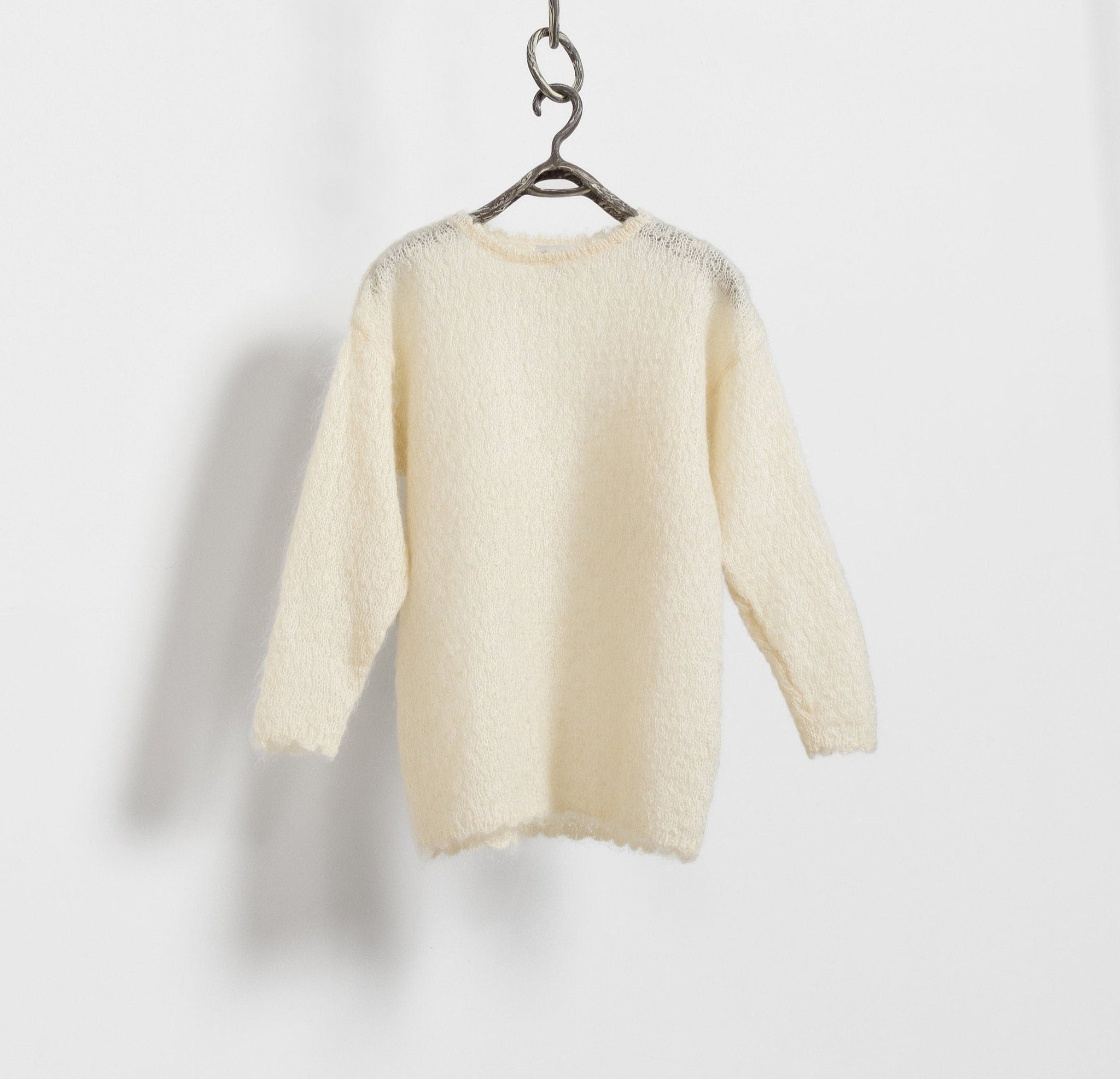 A vintage 90's cream mohair-blend pullover sweater hanging on a hook against a white door, displaying its loose, open knit and crew neck design.