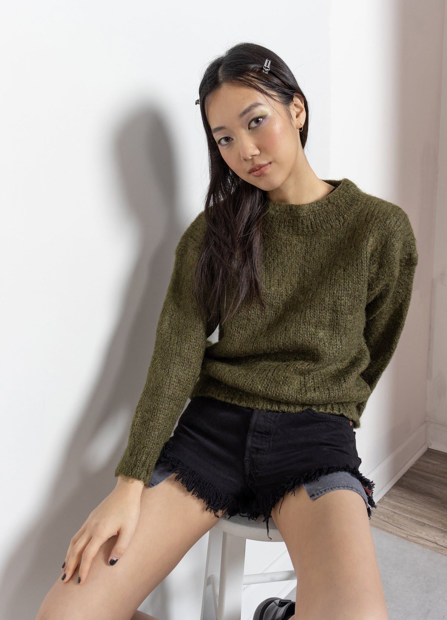 A woman sitting on a chair wearing a vintage 90's olive green mohair-blend mock-neck pullover sweater and black denim shorts.