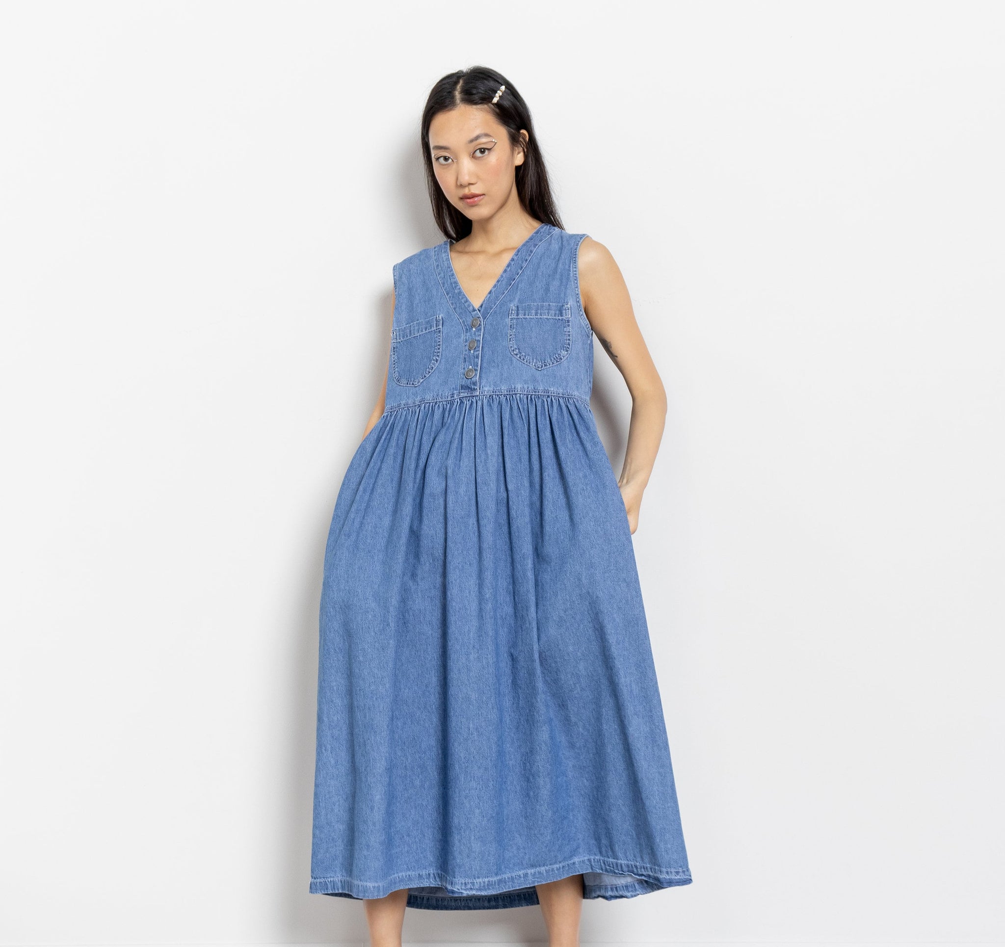 A woman modeling a sleeveless, mid-length denim dress with metal buttons at the bust and two front breast pockets.
