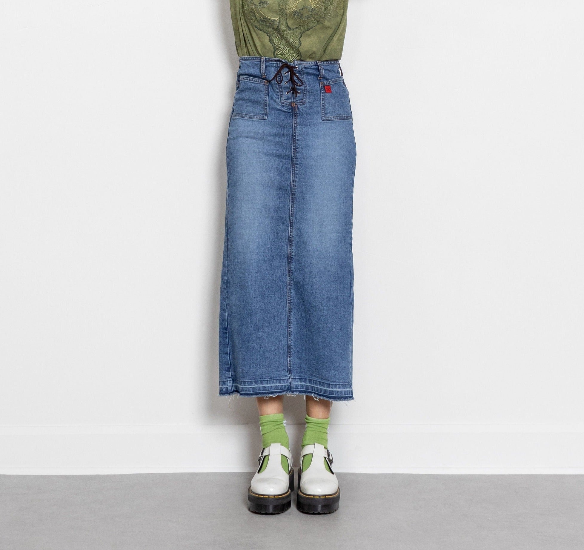 A person wearing a vintage Y2K crazy fade denim low rise pencil skirt with a corset style waistband, in a green-toned photo. The skirt has lighter faded patches and is paired with white sneakers.