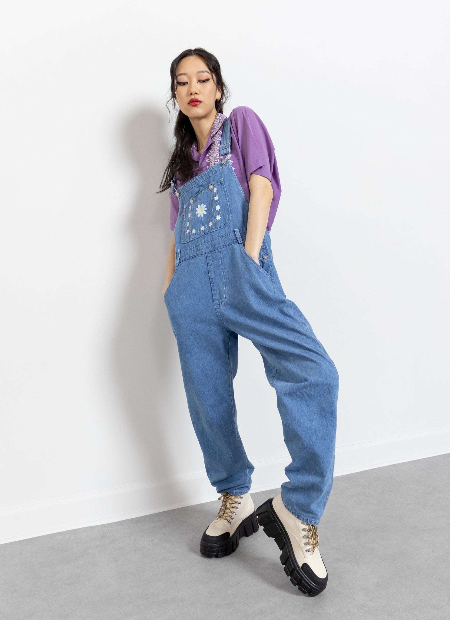 A person wearing vintage denim overalls with embroidered daisies on the front pouch and back, featuring side and back pockets, and silver-tone grommets. The overalls are presented in a well-lit indoor setting.