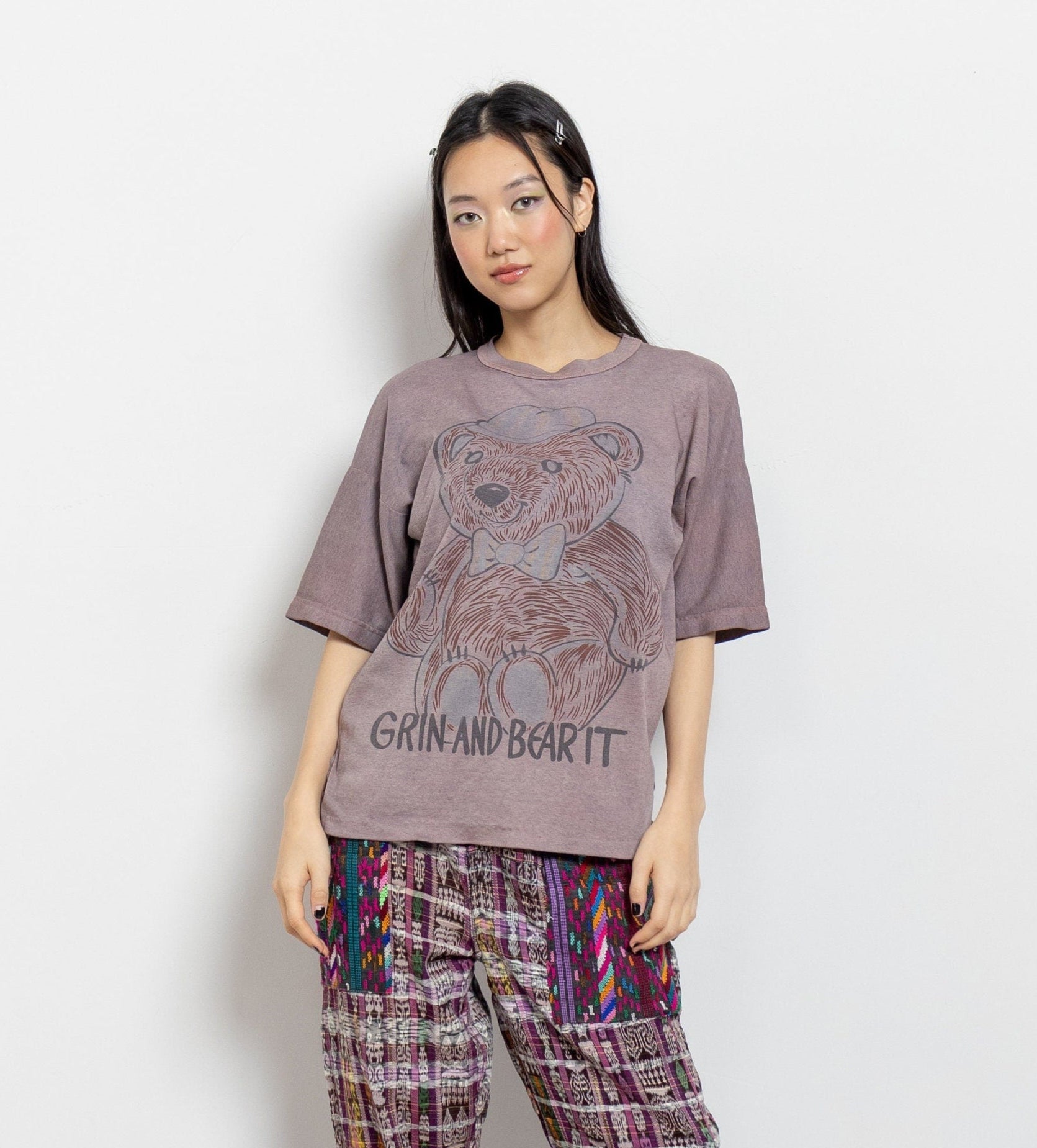 A woman modeling a vintage oversize brown t-shirt with a graphic of a bear wearing a bow tie and the text 'GRIN AND BEAR IT'. The t-shirt is faded tie-dye pale brown with a painterly style.