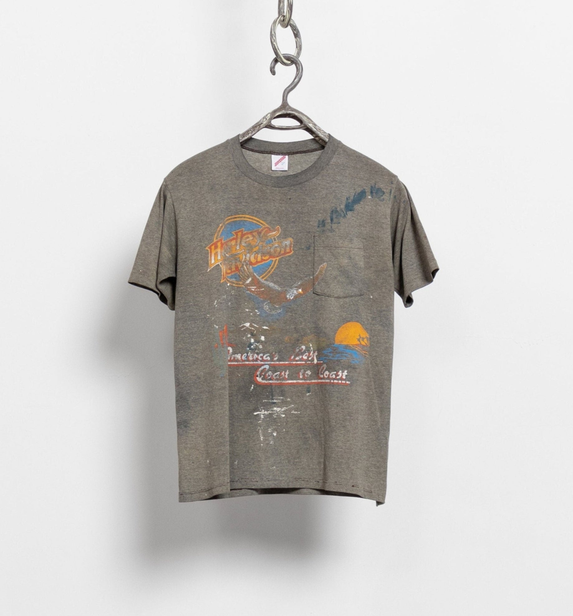 A vintage 80's Harley Davidson short sleeve tee hanging on a hanger, featuring front pocket graphic and a back graphic of a Harley dealer.